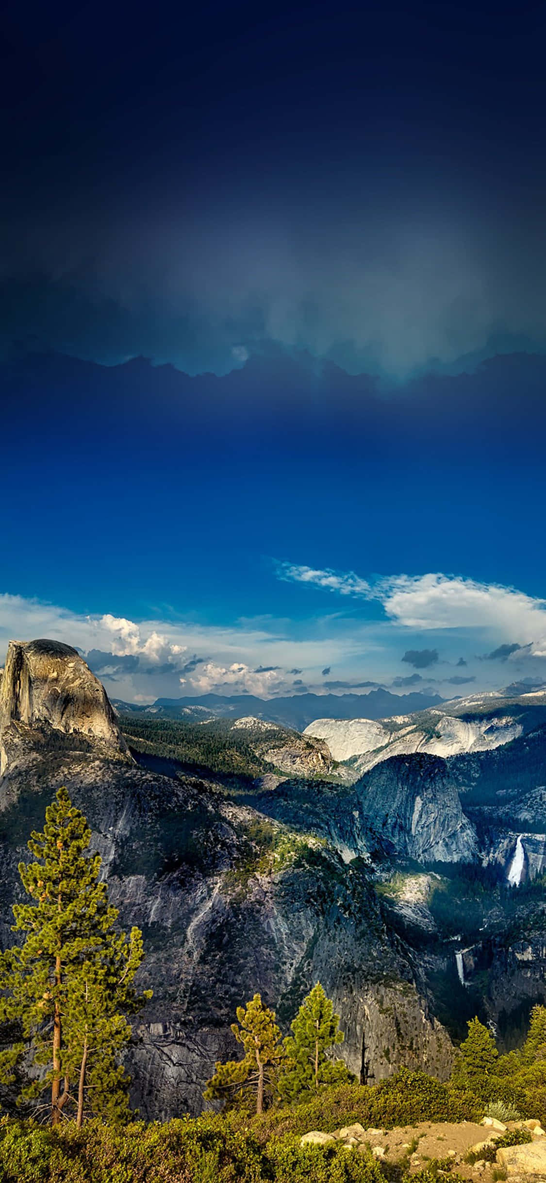 iPhone X Nature 桌布 壁紙 — 戲劇性的山景，Half Dome 在深藍色天空下，散落著雲朵，前景有松樹。