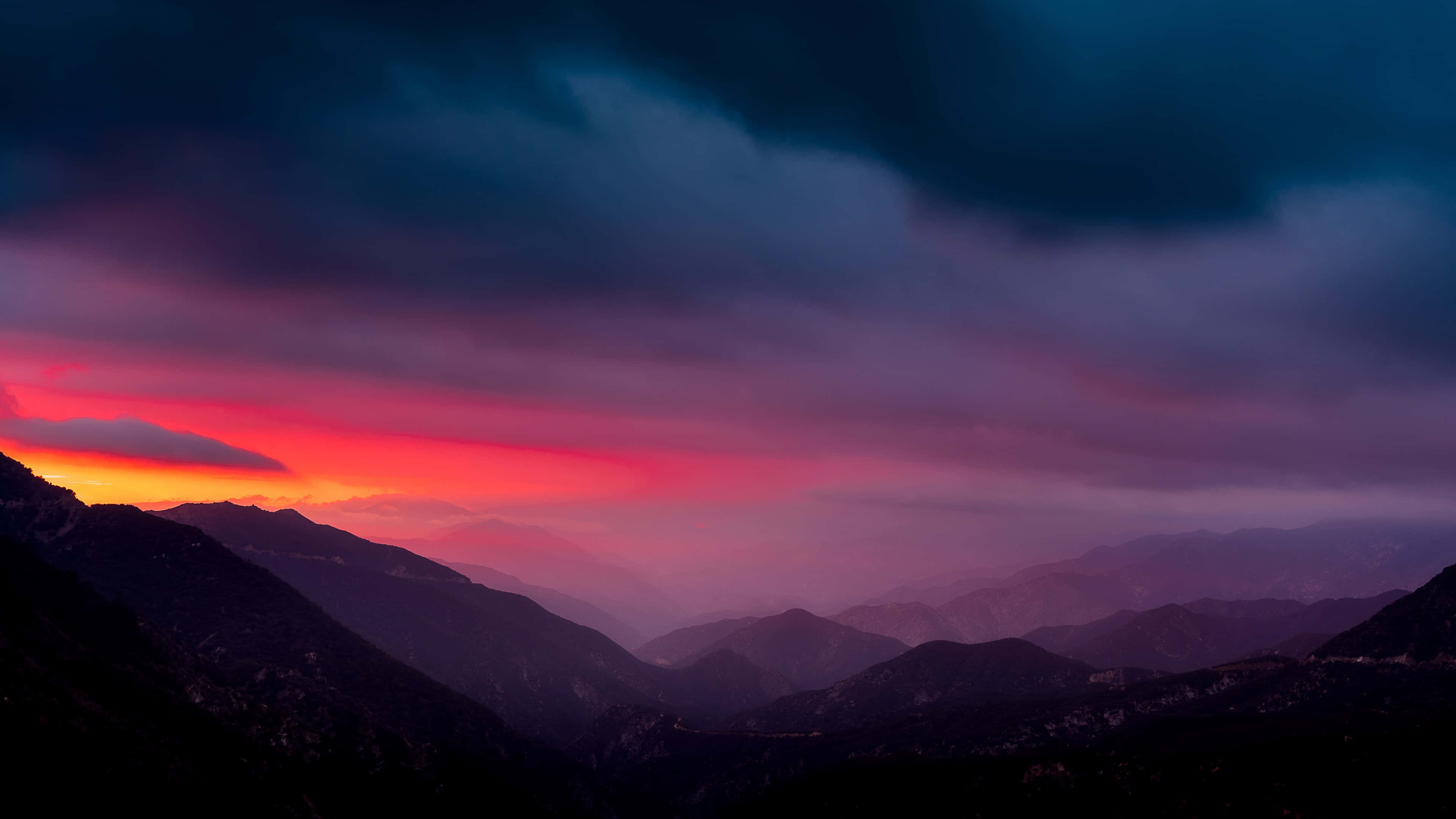Colorful Sky 桌布 壁紙 — 戲劇性的山脈輪廓映襯著火燒般的日落天空，天空中有深紫色和橙色的雲彩。