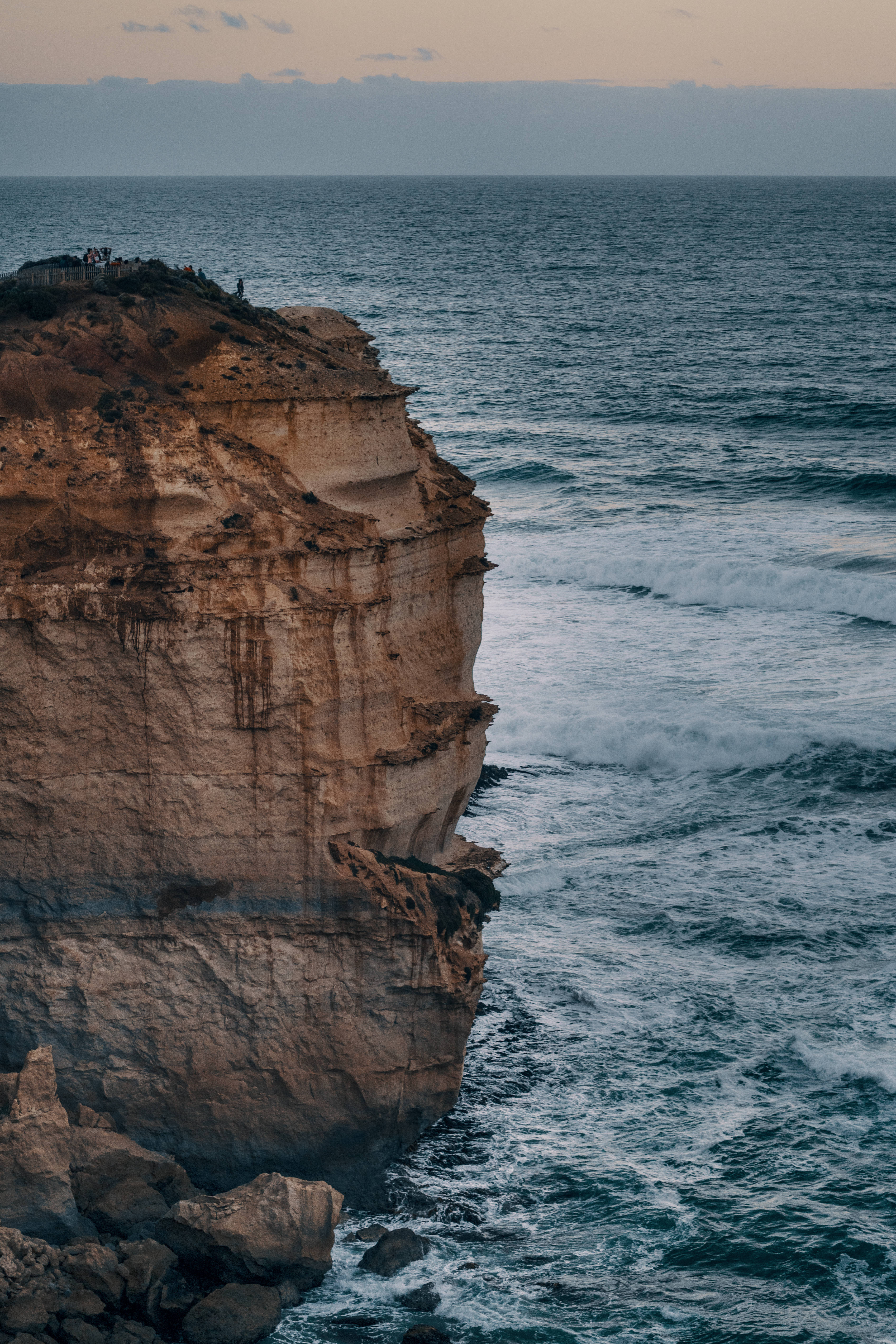 australia iPhone 桌布 壁紙 — 戲劇性的懸崖面在柔和暮色天空下與洶湧的海浪相遇，邊緣有遠處的人影。