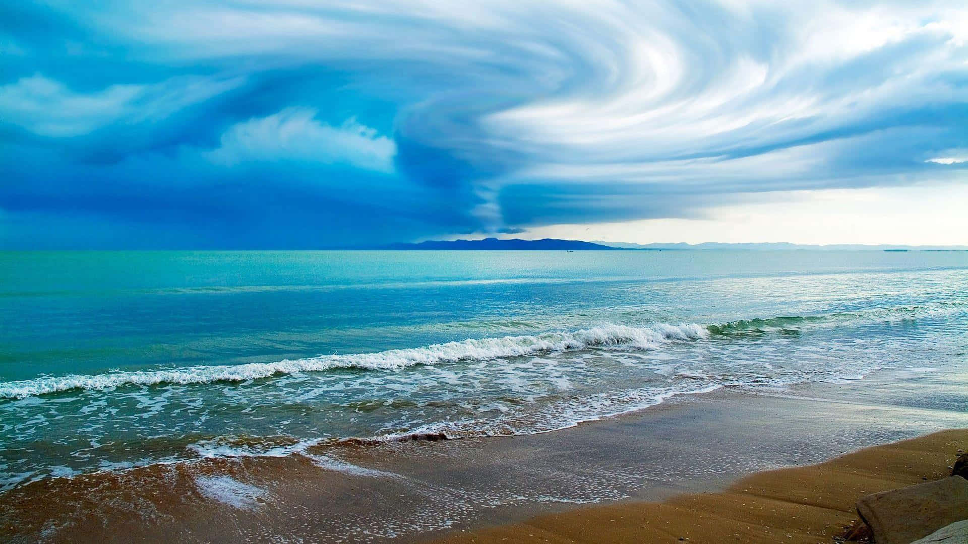 Beach Desktop 桌布 壁紙 — 戲劇性的旋轉暴風雲籠罩在碧綠海洋與金色沙灘交匯處的上空。