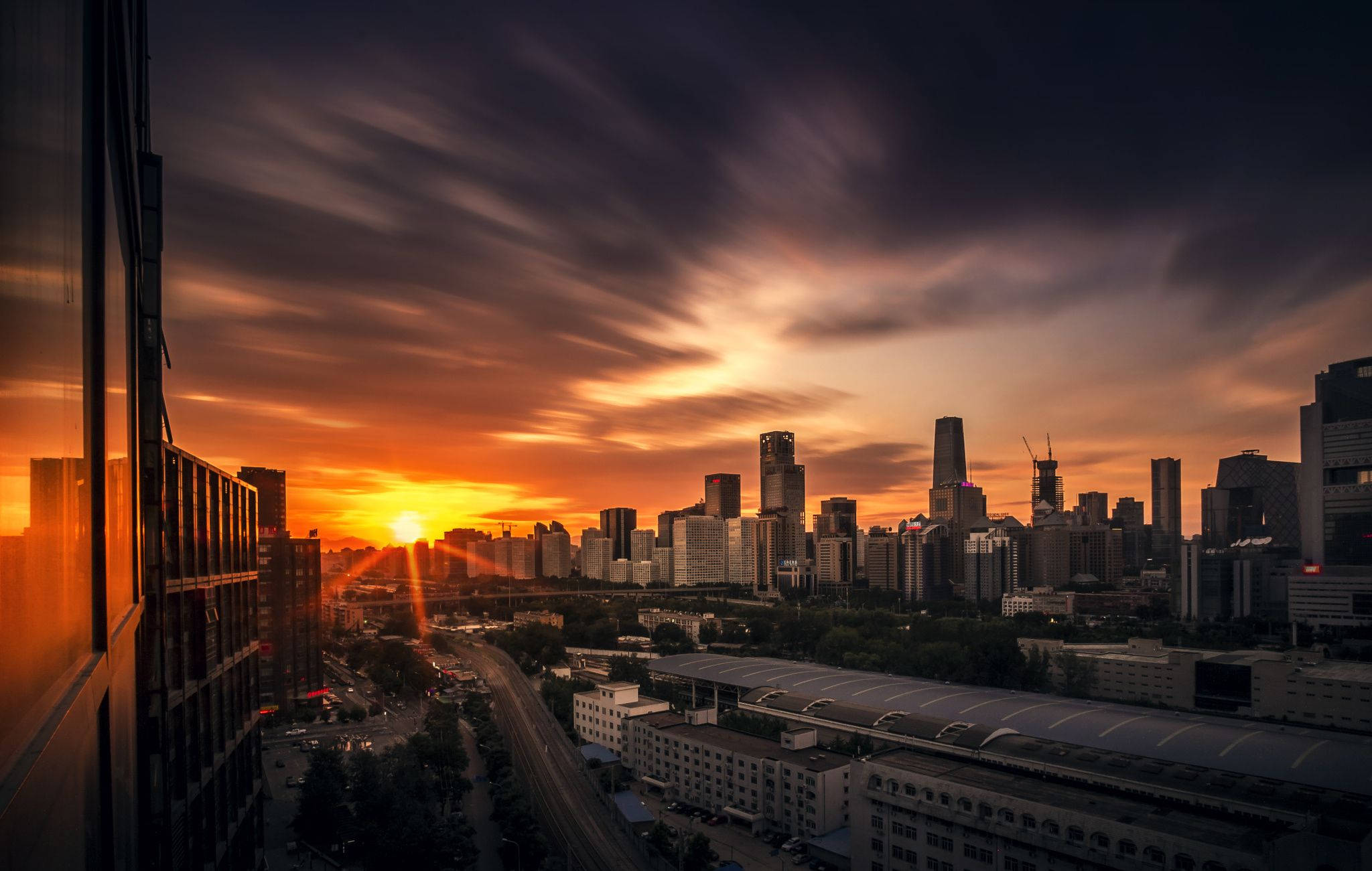 Beijing 桌布 壁紙 — 戲劇性的日落以火橙色和深紫色調沐浴現代城市景觀，雲彩劃過天空。