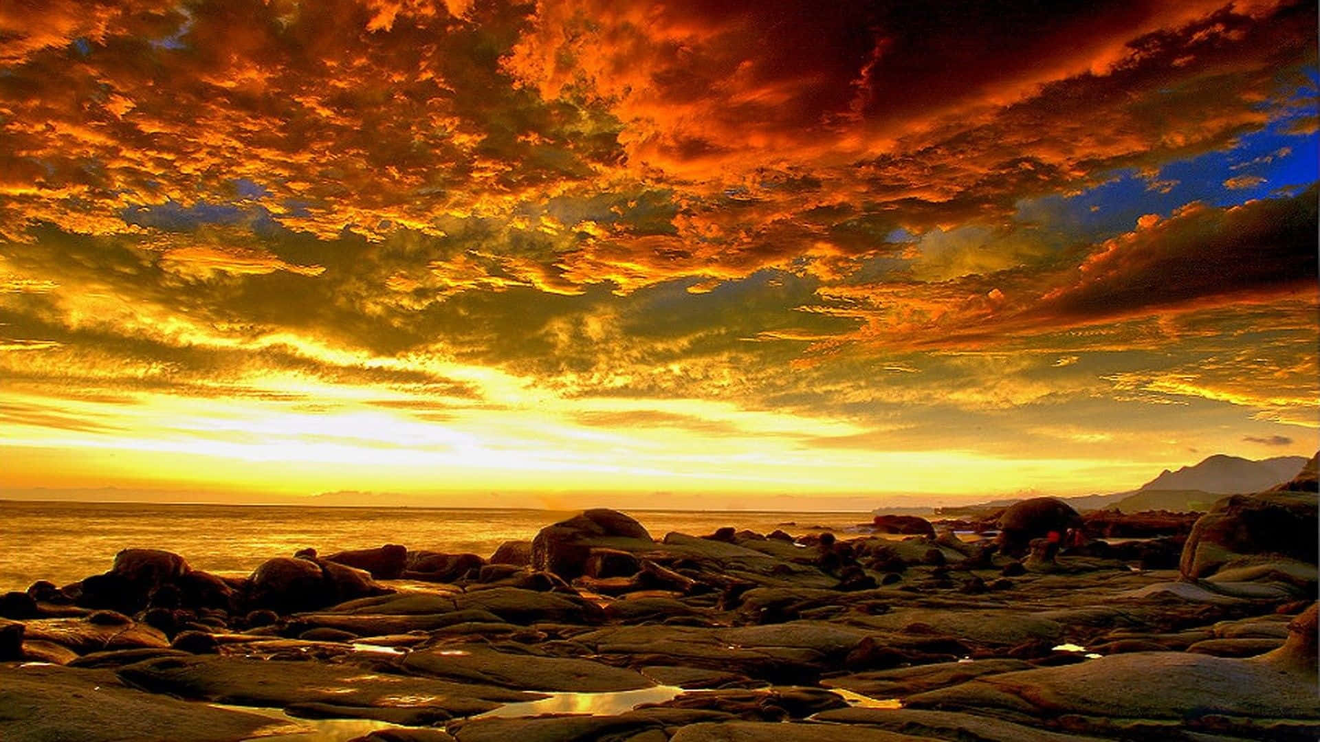 Beautiful Sky 桌布 壁紙 — 戲劇性的日落將天空染成火橙色和金色，映照在岩石海岸線和遠處的山巒之上。