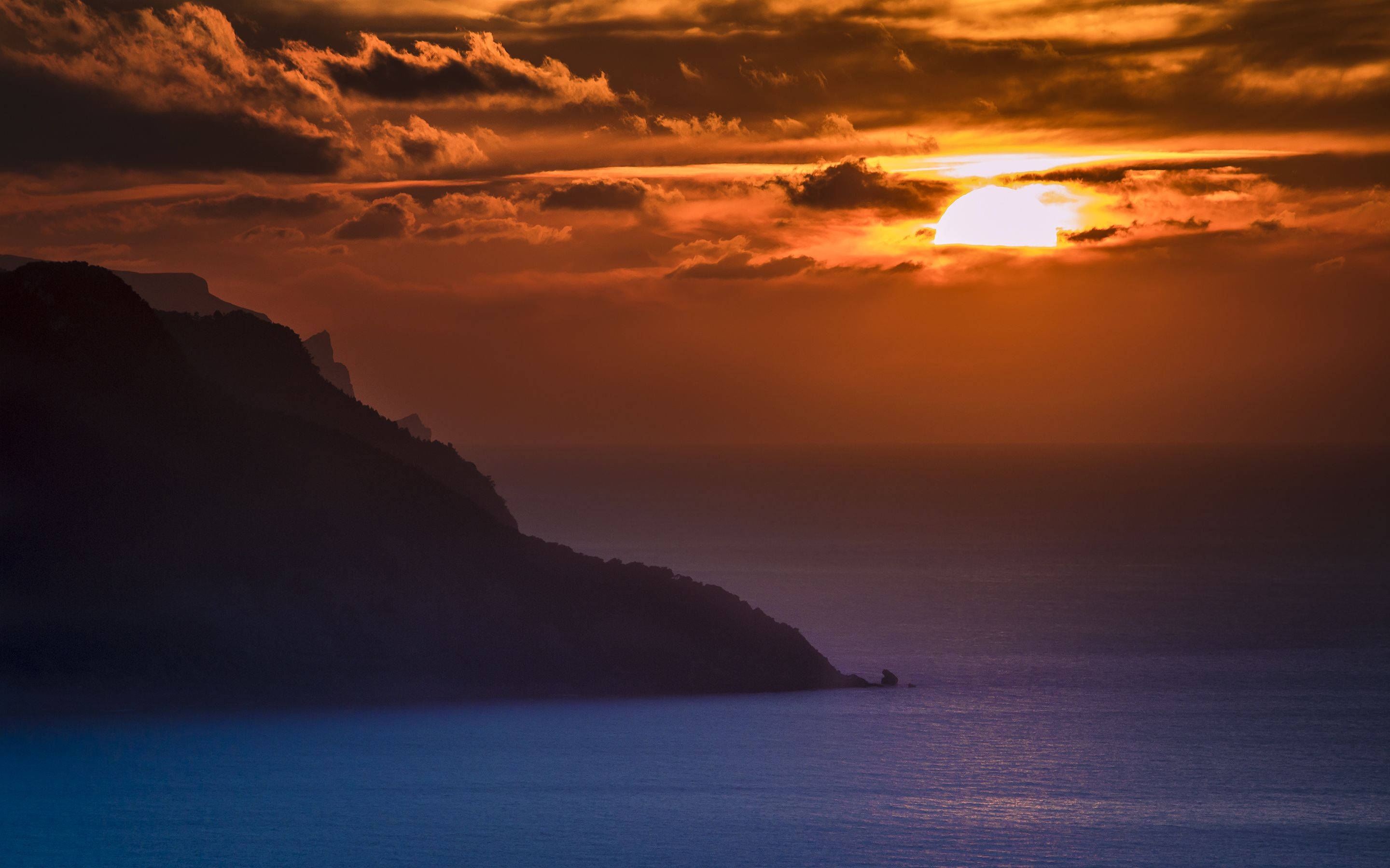 海景 桌布 壁紙 — 戲劇性的日落將天空染成火熱的橙色，陽光穿過雲層照射在黑暗的懸崖和寧靜的藍色海洋上。
