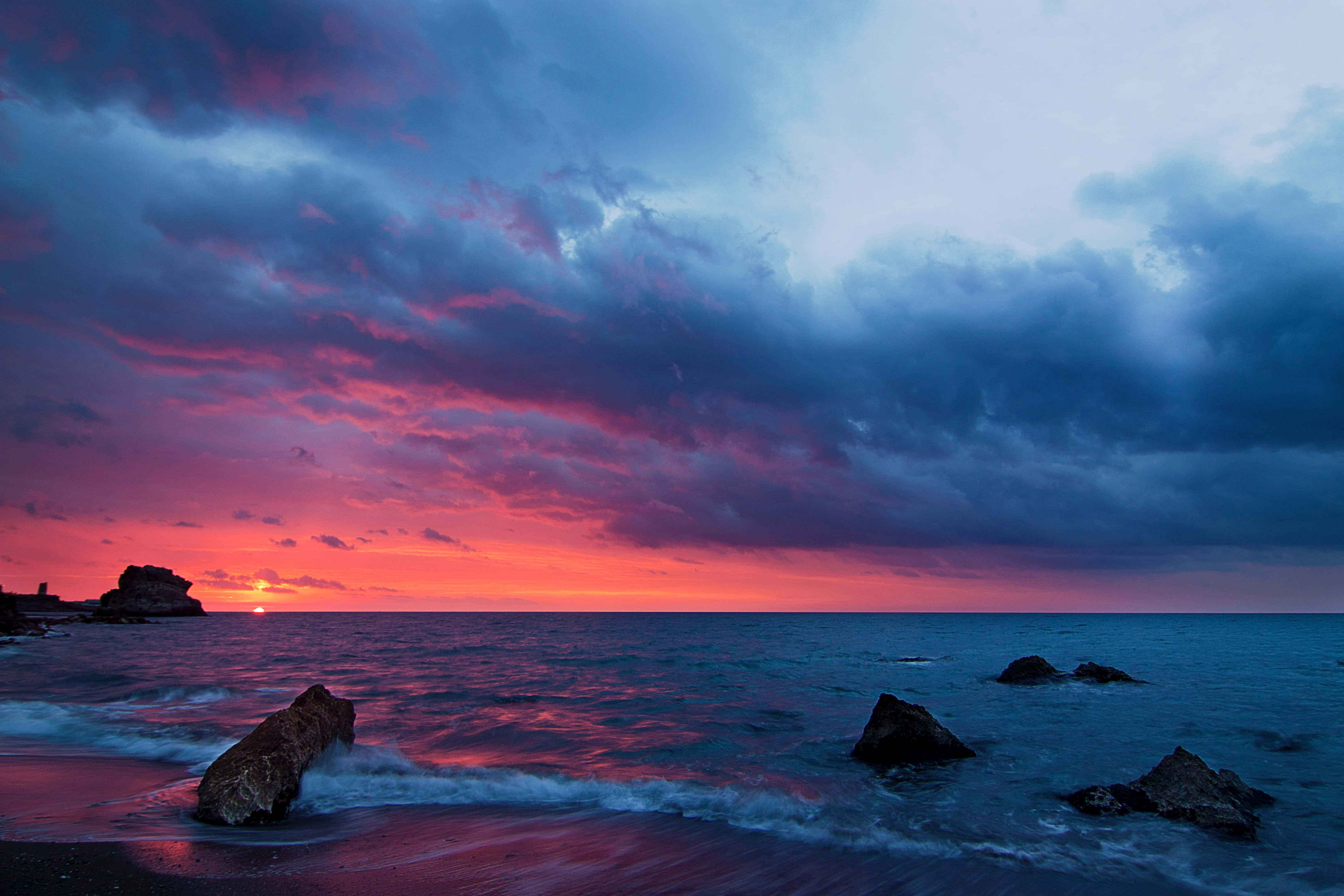 Colorful Sky 桌布 壁紙 — 戲劇性的日落將天空染成火熱的橙色和深紫色，海浪拍打著寧靜海岸線上的黑色岩石。