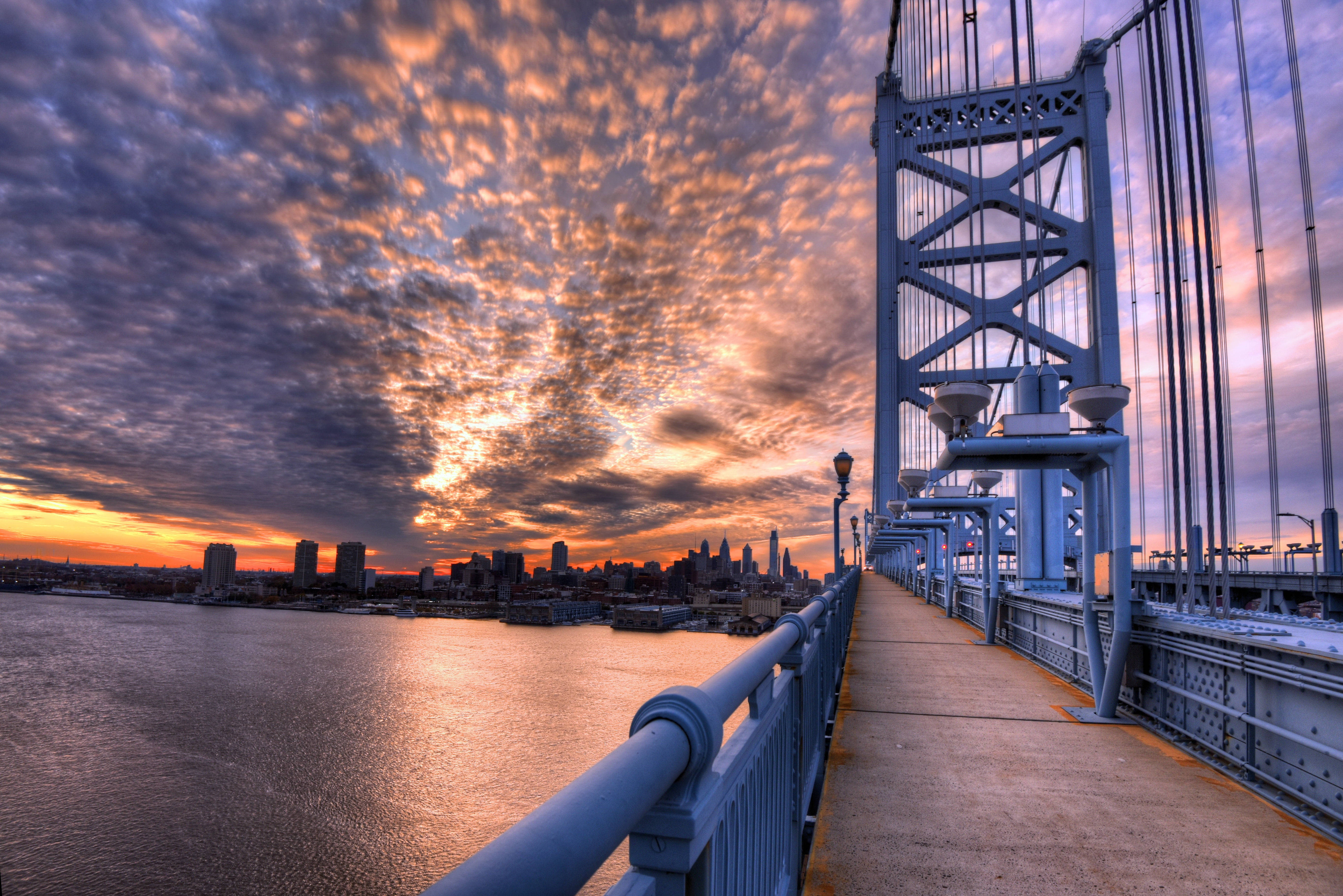 Philadelphia 桌布 壁紙 — 戲劇性的日落將天空染成火熱的橙色和紫色，鋼橋俯瞰城市天際線和水面。