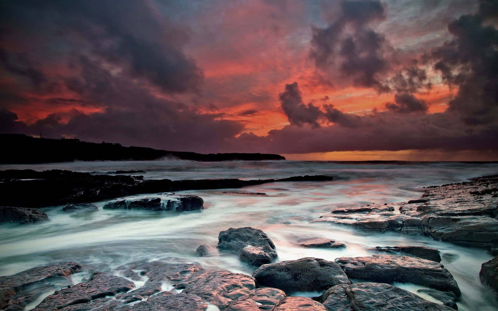 Photography Desktop 桌布 壁紙 — 戲劇性的日落將天空染成火紅和紫色，海浪平穩地流過深色火山岩。
