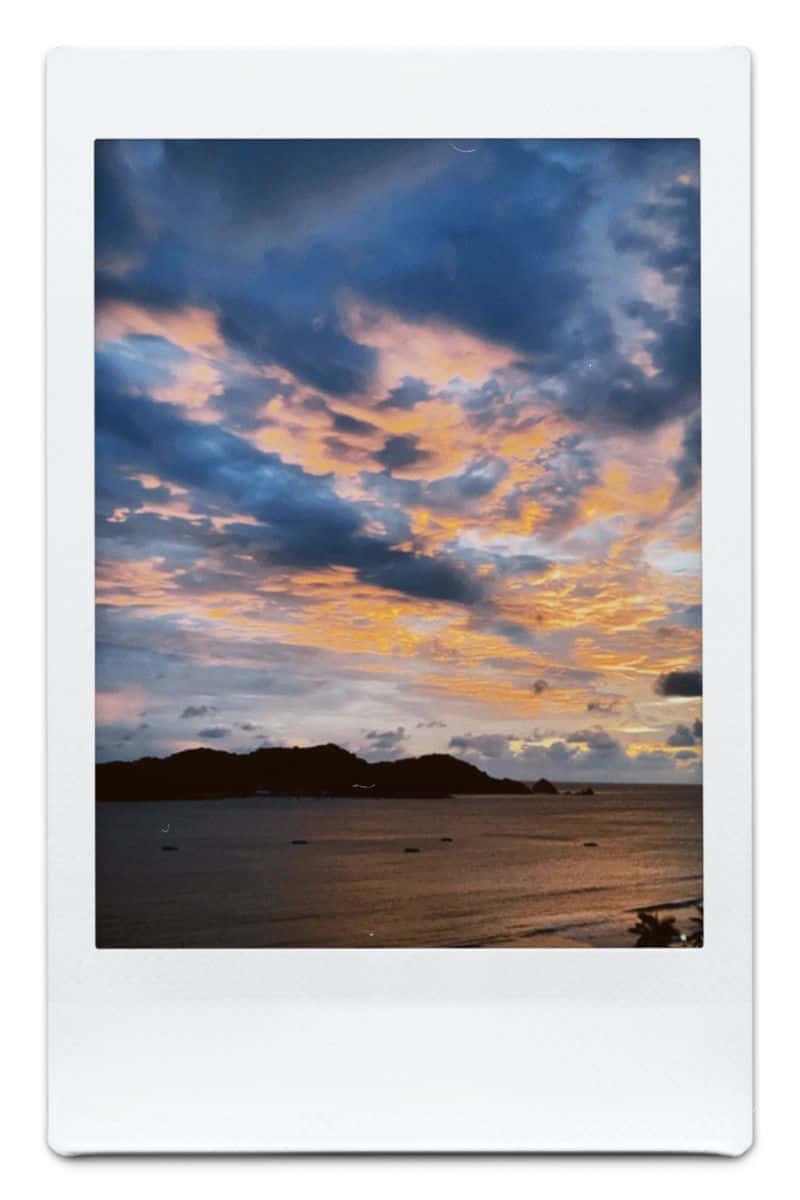 Aesthetic Polaroid 桌布 壁紙 — 戲劇性的日落將雲彩染成火橙色和深藍色，映照在平靜的海面上，遠處有山丘和船隻。