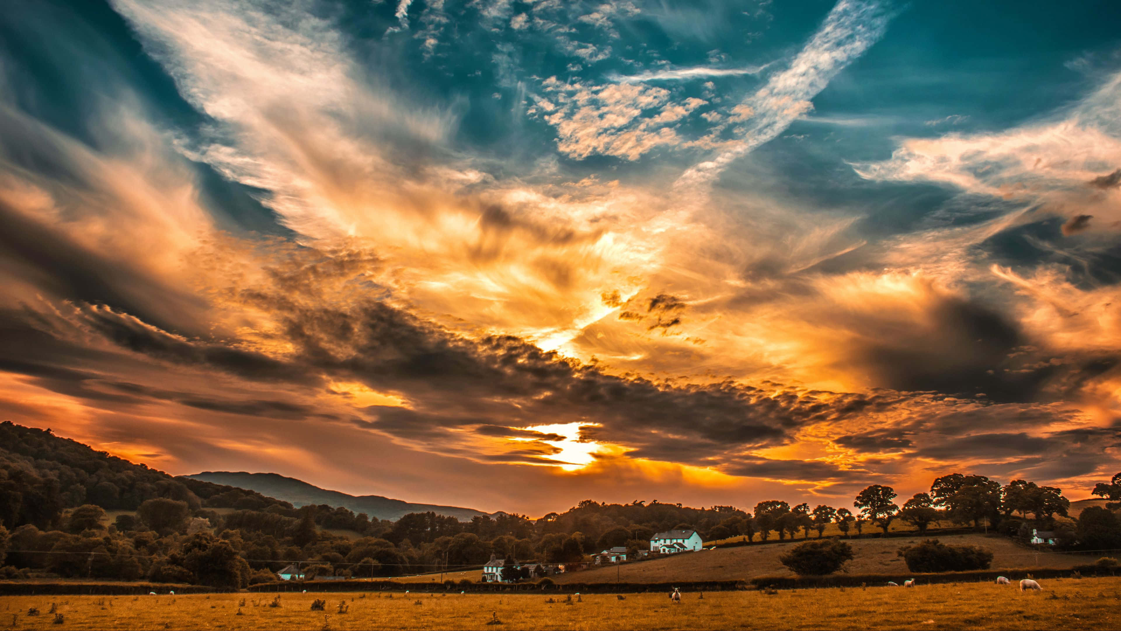 Colorful Sky 桌布 壁紙 — 戲劇性的日落將雲彩染成火橙色和藍綠色，映照在點綴著樹木和遠處農舍的起伏山丘上。