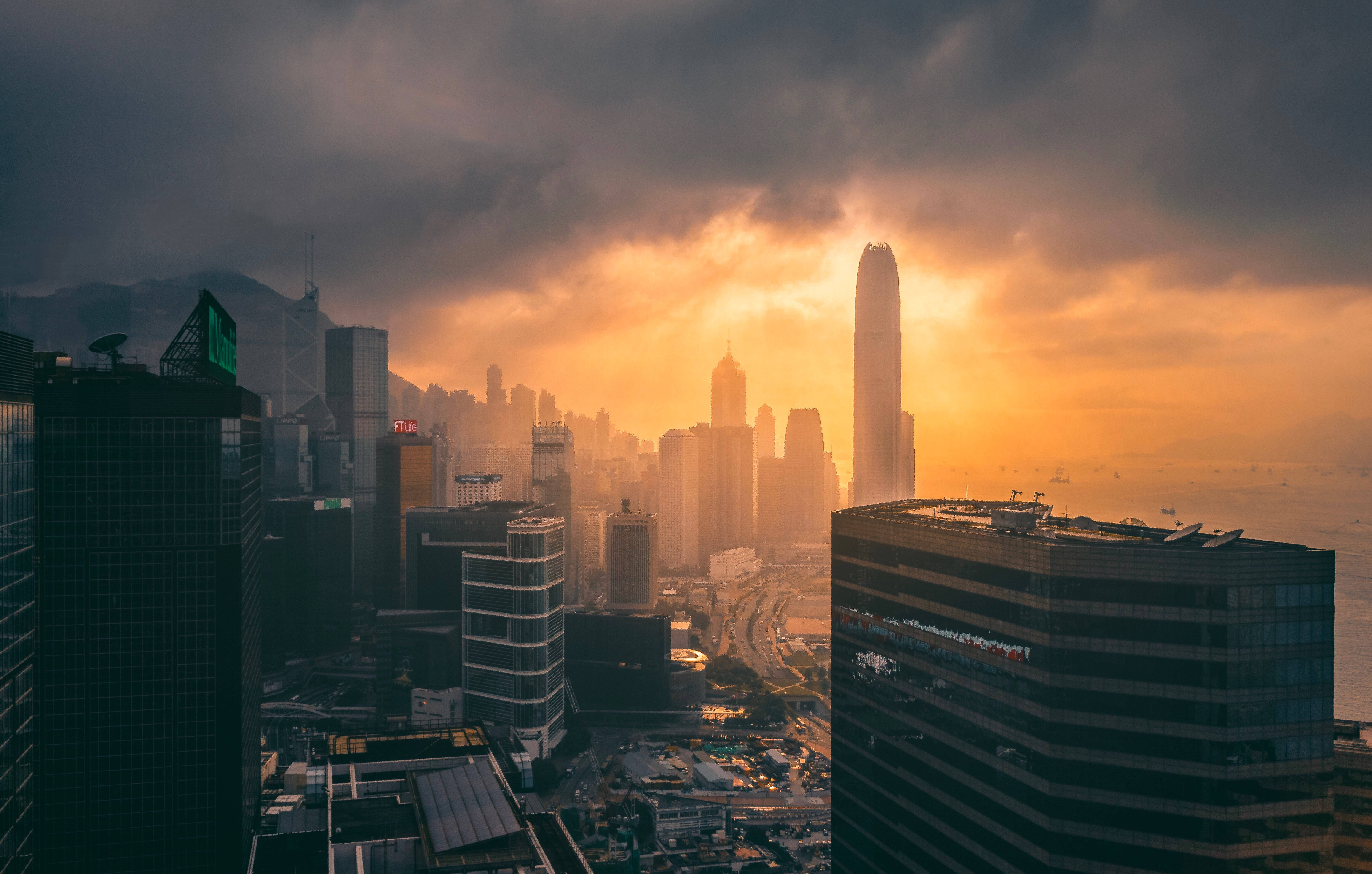 城市 桌布 壁紙 — 戲劇性的日落將香港的天際線沐浴在金色光芒中，烏雲籠罩著高聳的摩天大樓和遠處的港口。