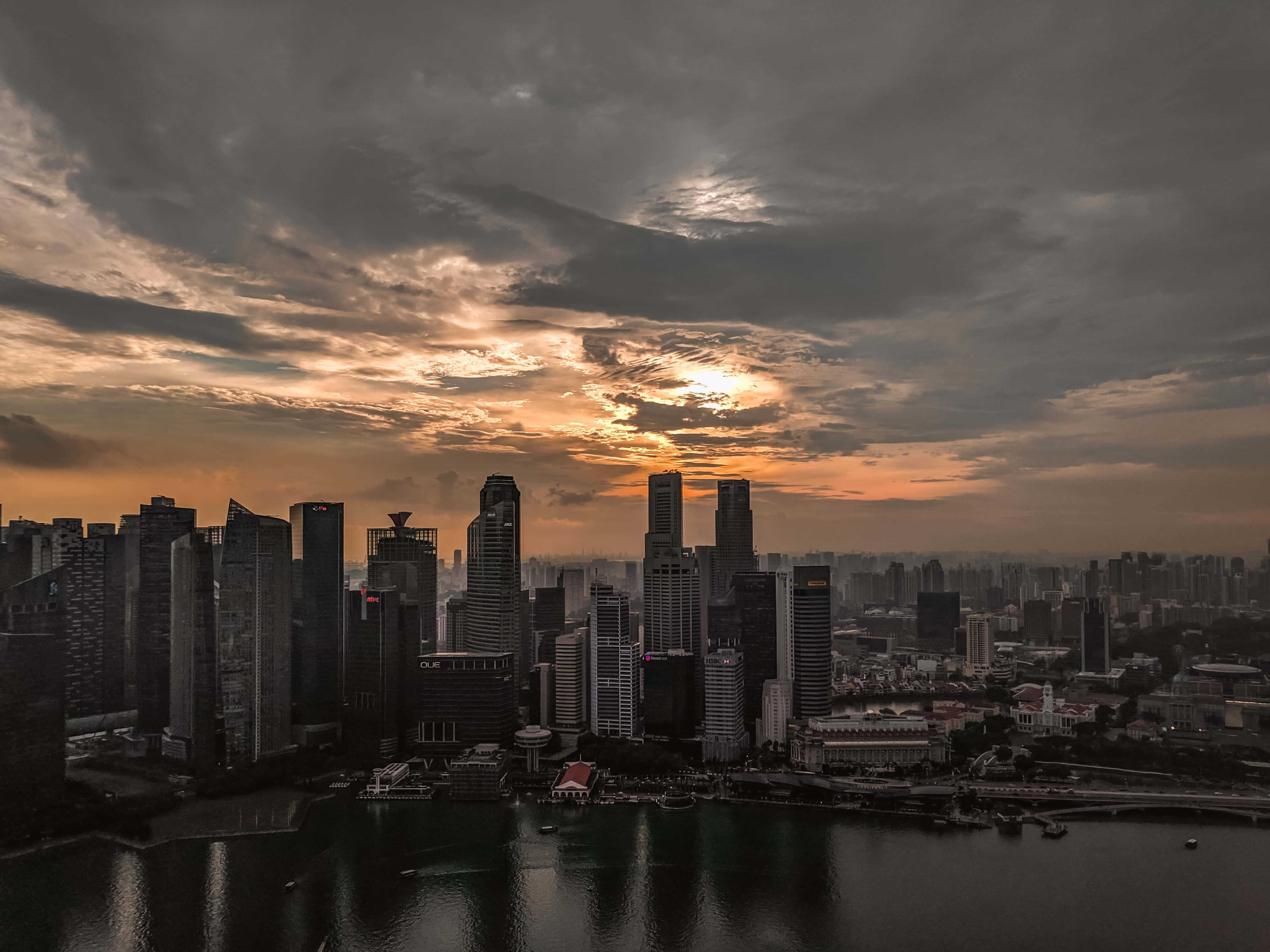 Twitter Header 桌布 壁紙 — 戲劇性的日落透過暴風雲灑下金色光芒，映照在新加坡高聳天際線在平靜水面的倒影上。