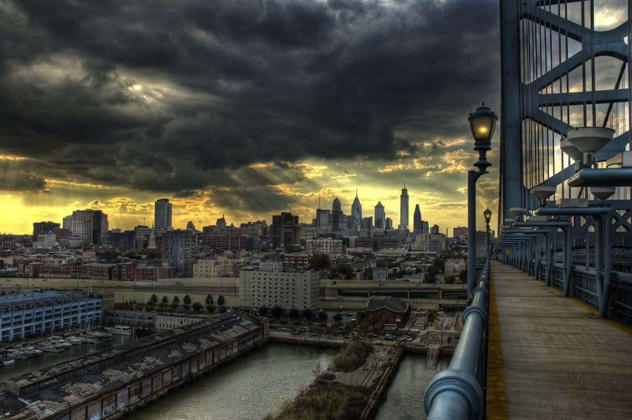 Philly 桌布 壁紙 — 戲劇性的暴風雲籠罩著日落時分的城市天際線，從一座帶有發光路燈的橋上觀看。