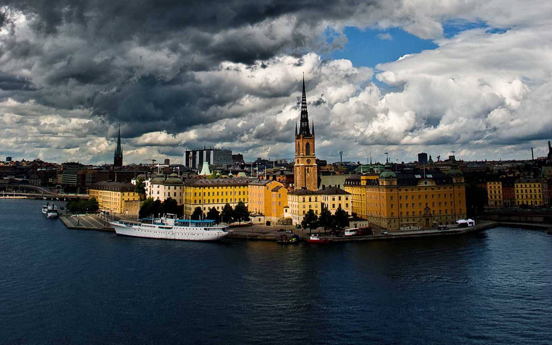 Sweden 桌布 壁紙 — 戲劇性的暴風雲籠罩著歷史悠久的濱水城市景觀，黃色建築和高聳的教堂尖塔矗立在黑暗的水邊。