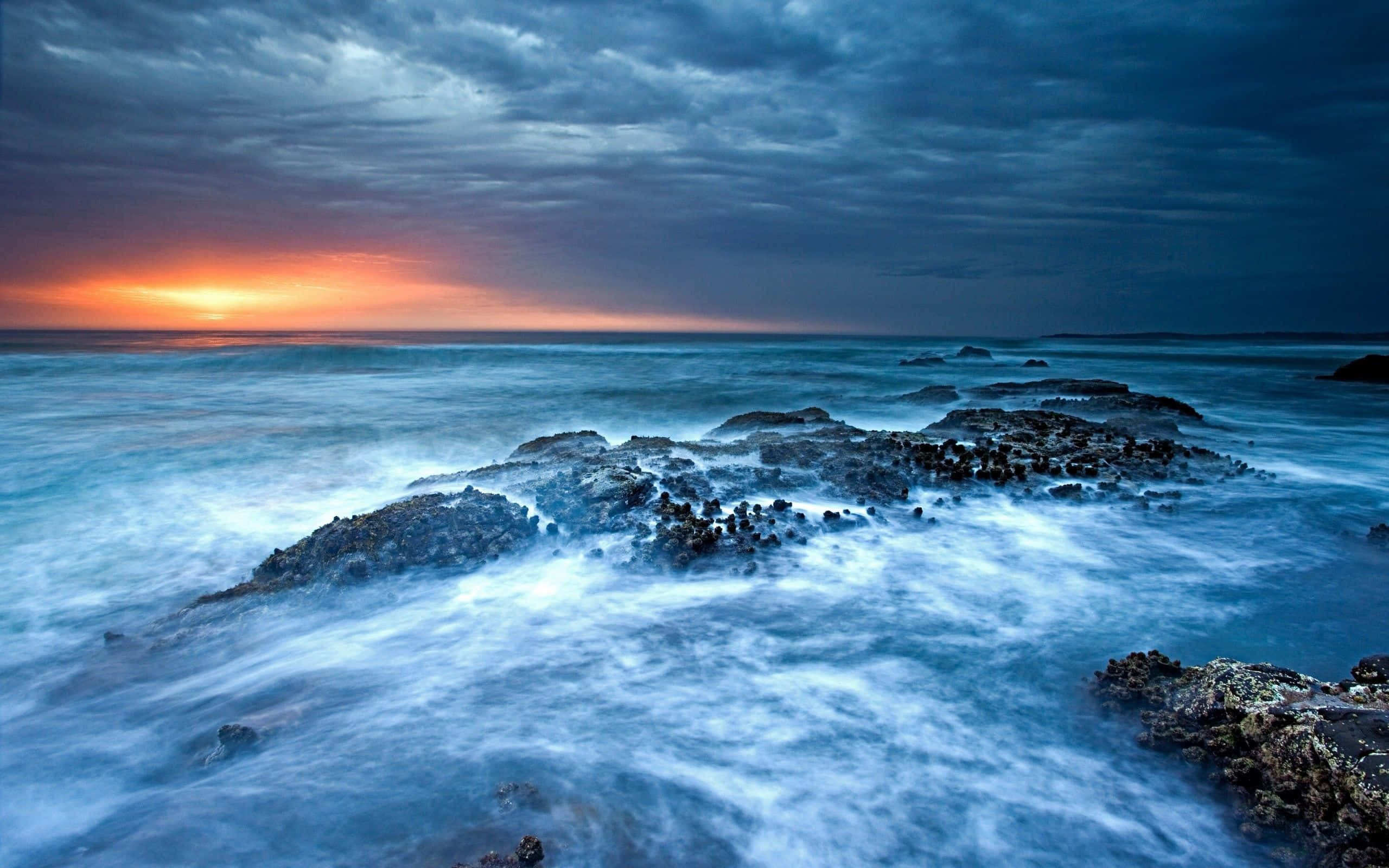 Ocean Waves 桌布 壁紙 — 戲劇性的海浪在暴風雨的天空下衝擊著黑暗的岩石，地平線上燃燒著夕陽的餘暉。