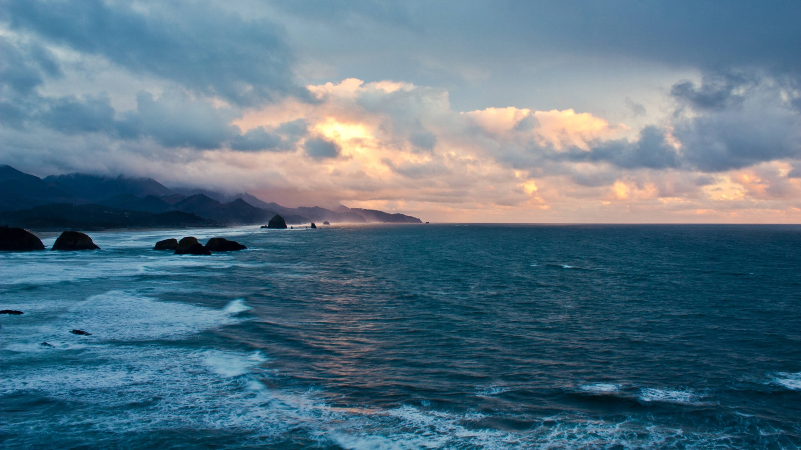 View 桌布 壁紙 — 戲劇性的海浪拍打著黑暗的岩石，在陰鬱的暮色天空下，伴隨著金色的雲彩。