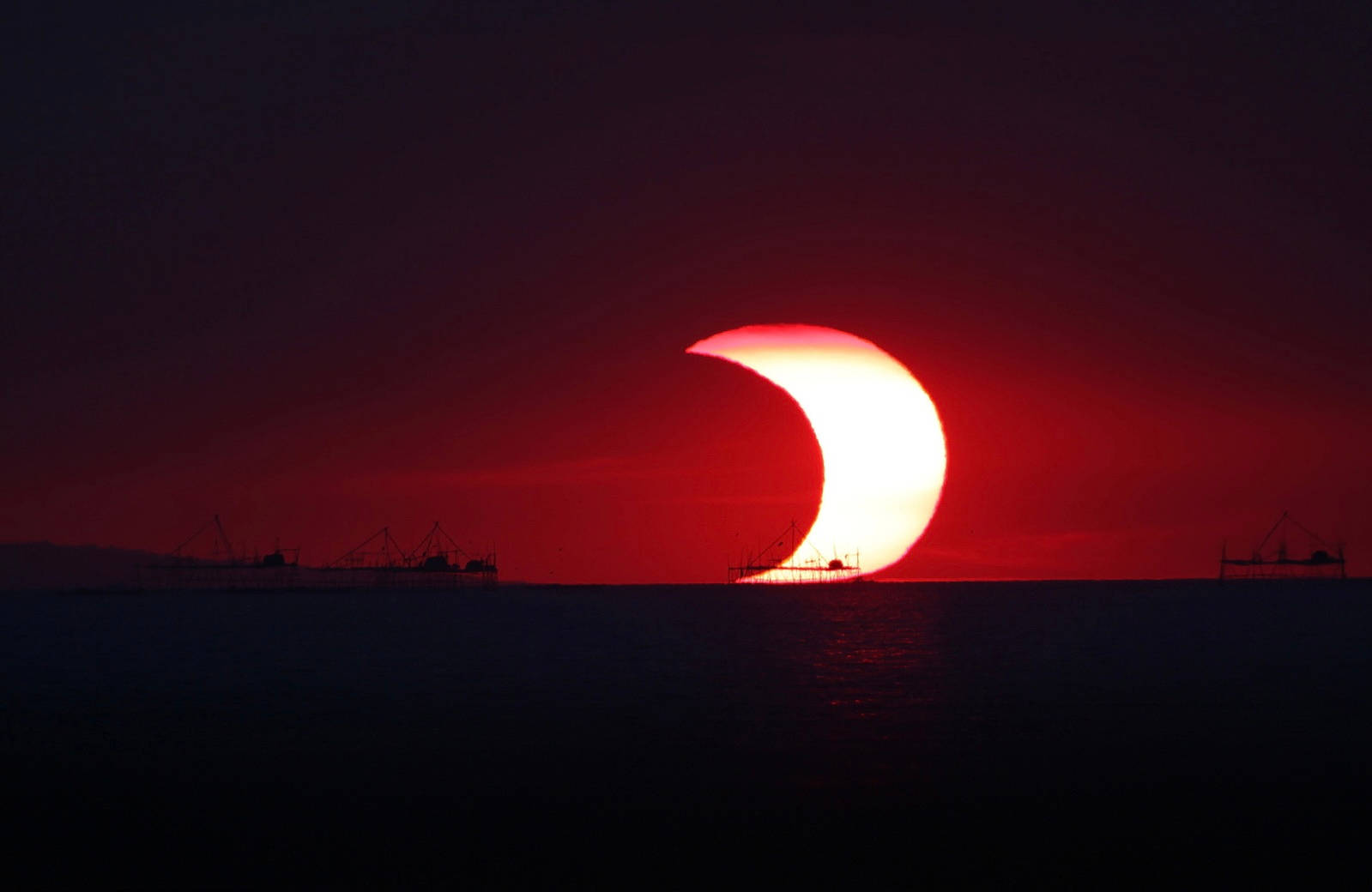 太陽與月亮 桌布 壁紙 — 戲劇性的紅色新月太陽在黑暗的海洋上發生食，漁具的剪影映襯在深紅色的天空中。