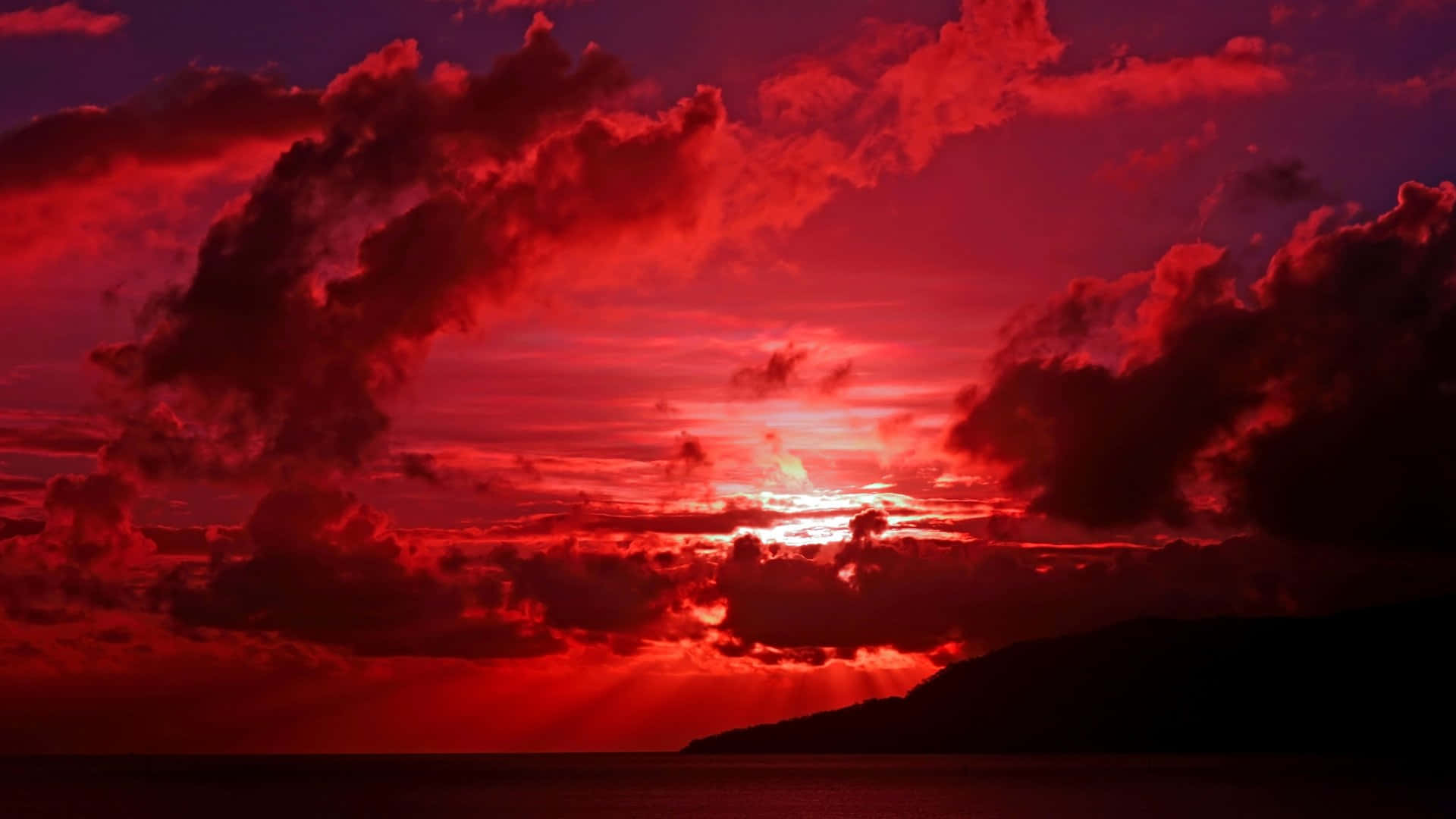 美學 桌布 壁紙 — 戲劇性的紅色日落，帶有火燒雲和光束，映照在黑暗的海面和山脈輪廓上。