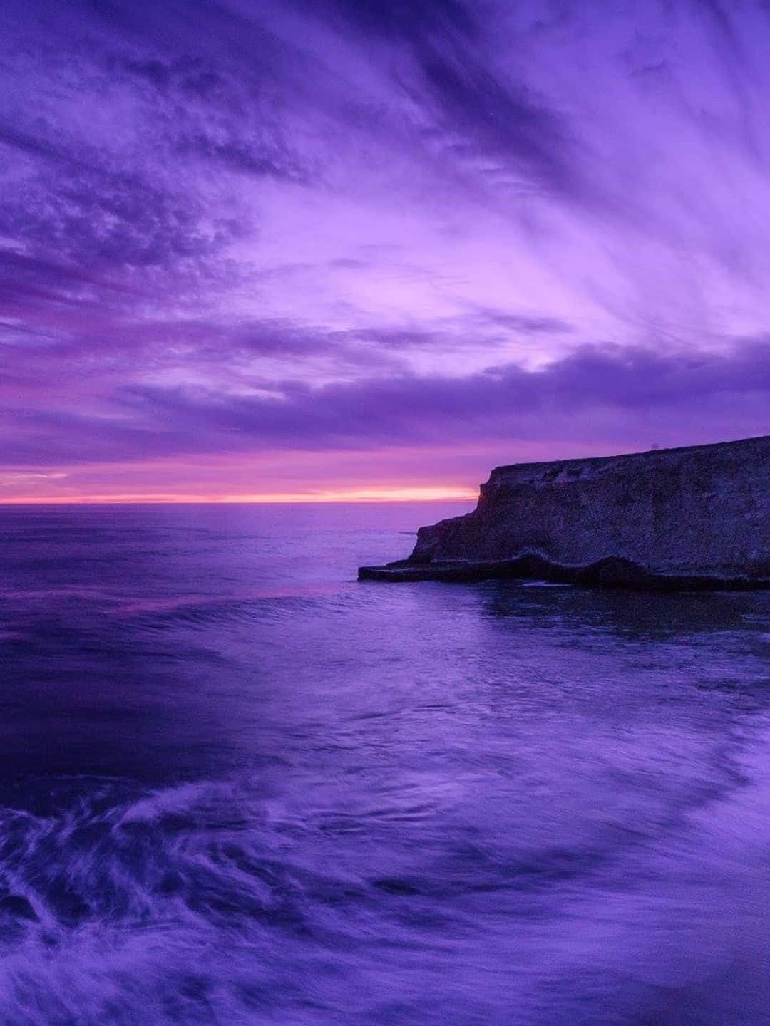 1440p Nature 桌布 壁紙 — 戲劇性的紫色日落為天空和海洋塗上色彩，海浪拍打崎嶇的懸崖。