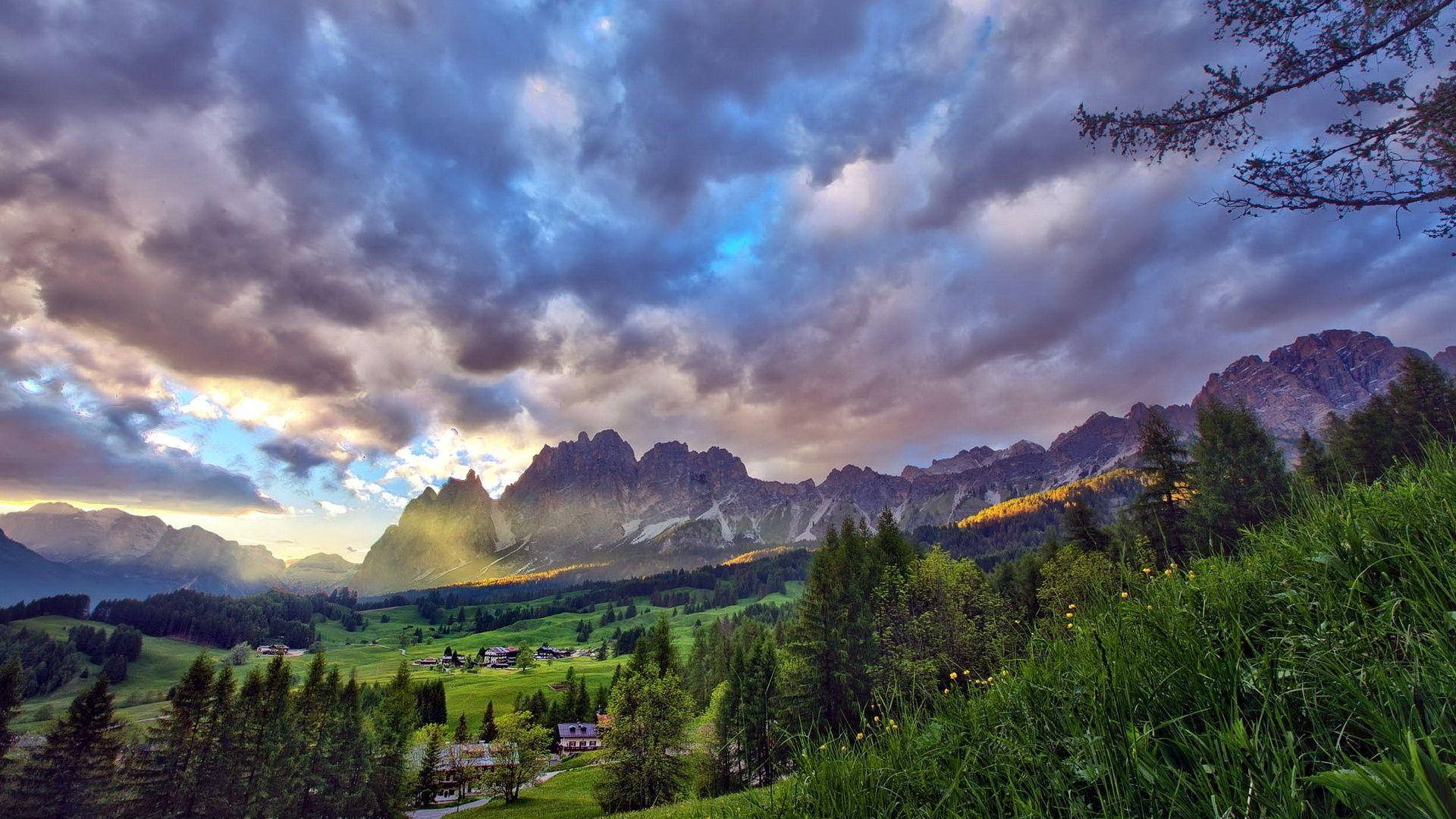 Beautiful Mountain View 桌布 壁紙 — 戲劇性的紫色雲彩在鋸齒狀的山峰上發光，金色光芒沐浴著一個有綠色草地和散落房屋的山谷。