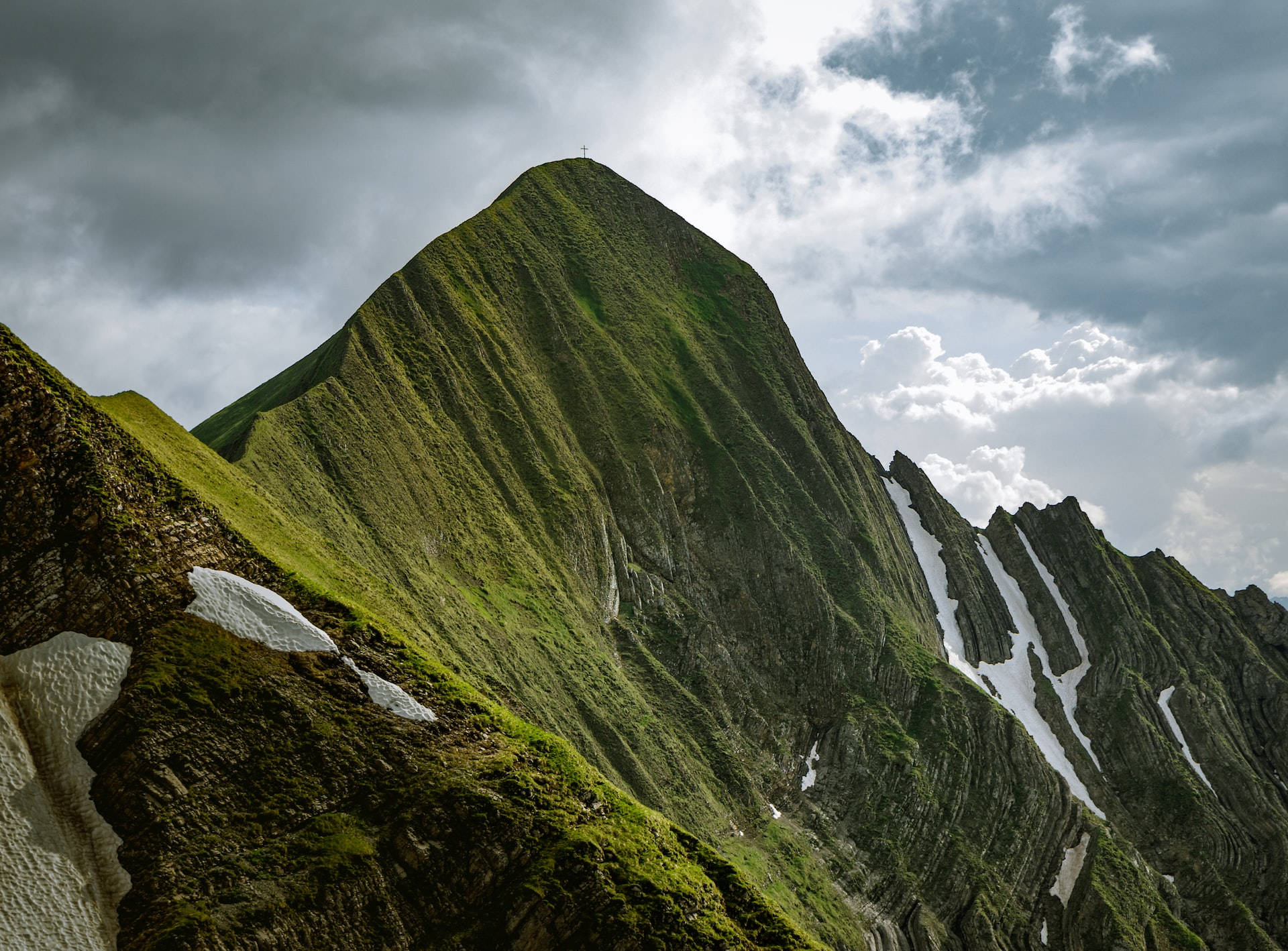 Mountain Landscape 桌布 壁紙 — 戲劇性的綠色山峰上殘留著雪塊，聳立在陰沉多雲的天空下。