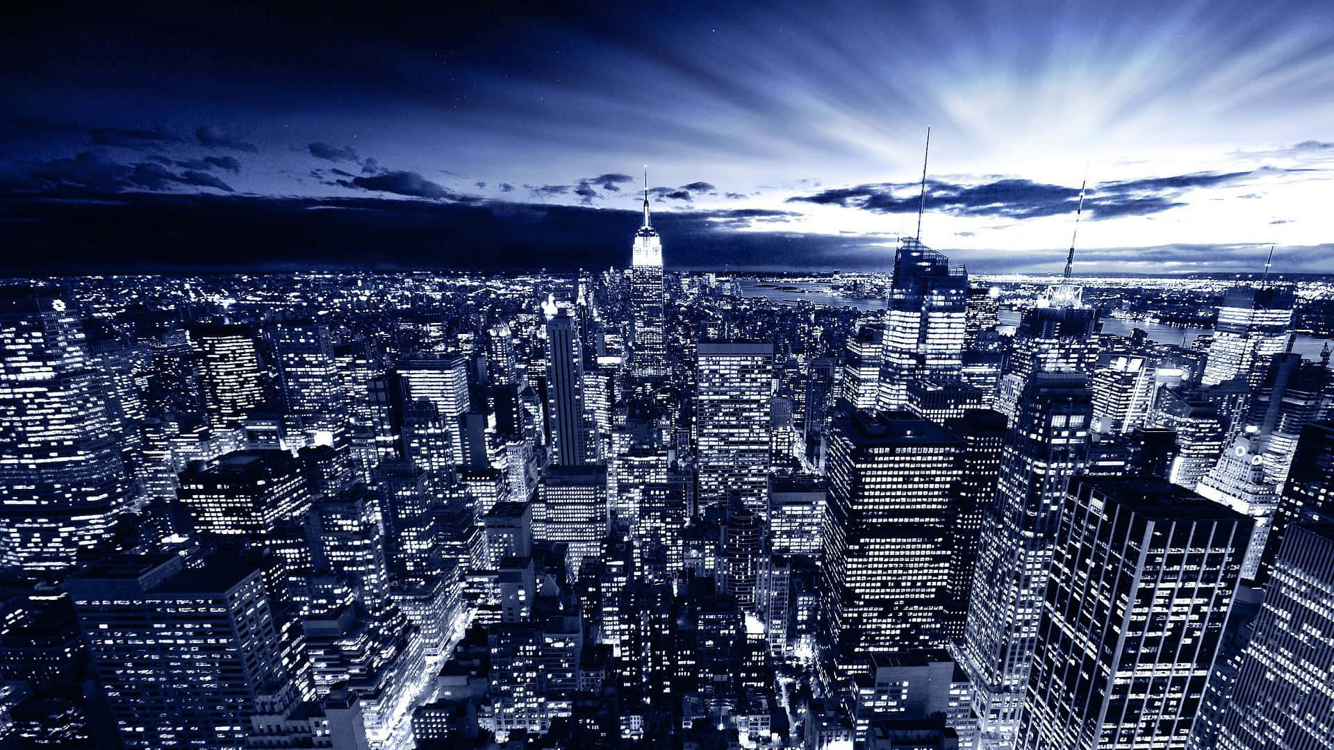 New York 桌布 壁紙 — 戲劇性的藍色調鳥瞰圖，展現夜晚廣闊的城市景觀，光芒四射的光束穿過雲層，照耀在高聳的摩天大樓之上。