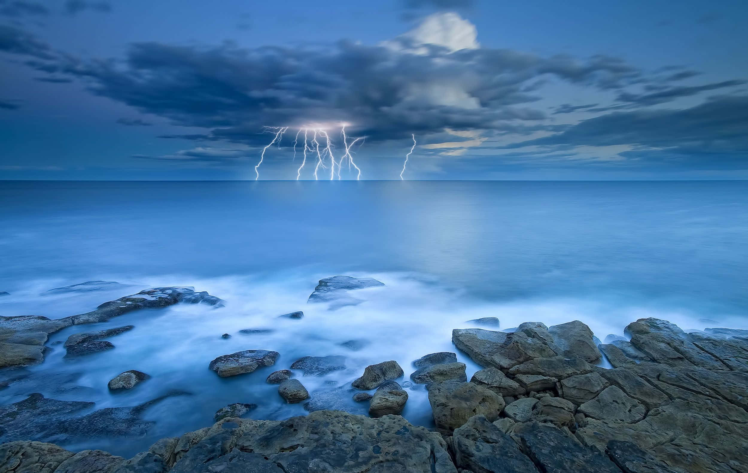 Ocean Storm 桌布 壁紙 — 戲劇性的閃電照亮了平靜海洋上空暴風雨的黃昏天空，前景是平靜的水面和參差不齊的岩石。