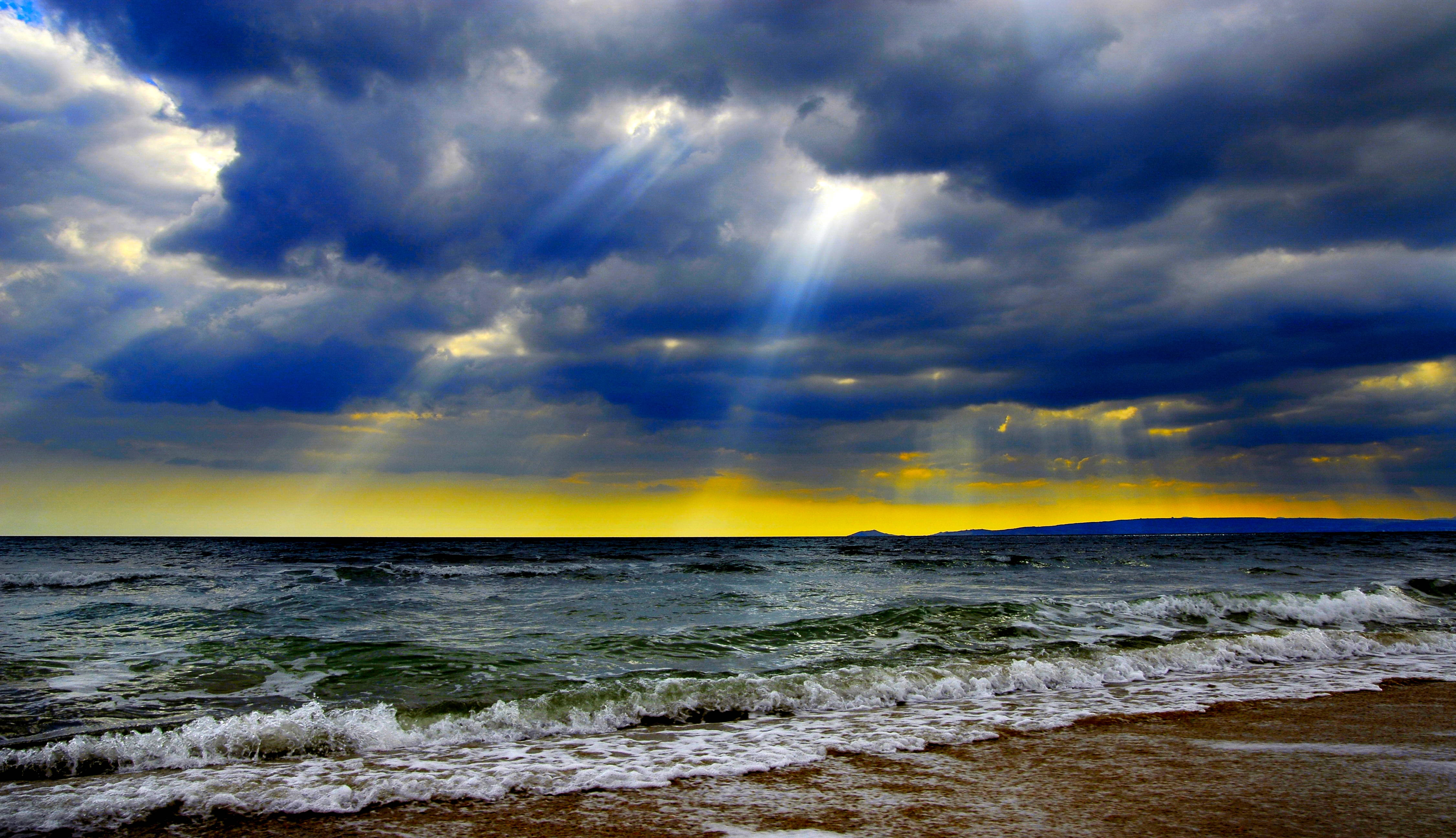 Sun Rays 桌布 壁紙 — 戲劇性的陽光穿透黑暗的暴風雲，照耀在波濤洶湧的大海與沙灘交會處。