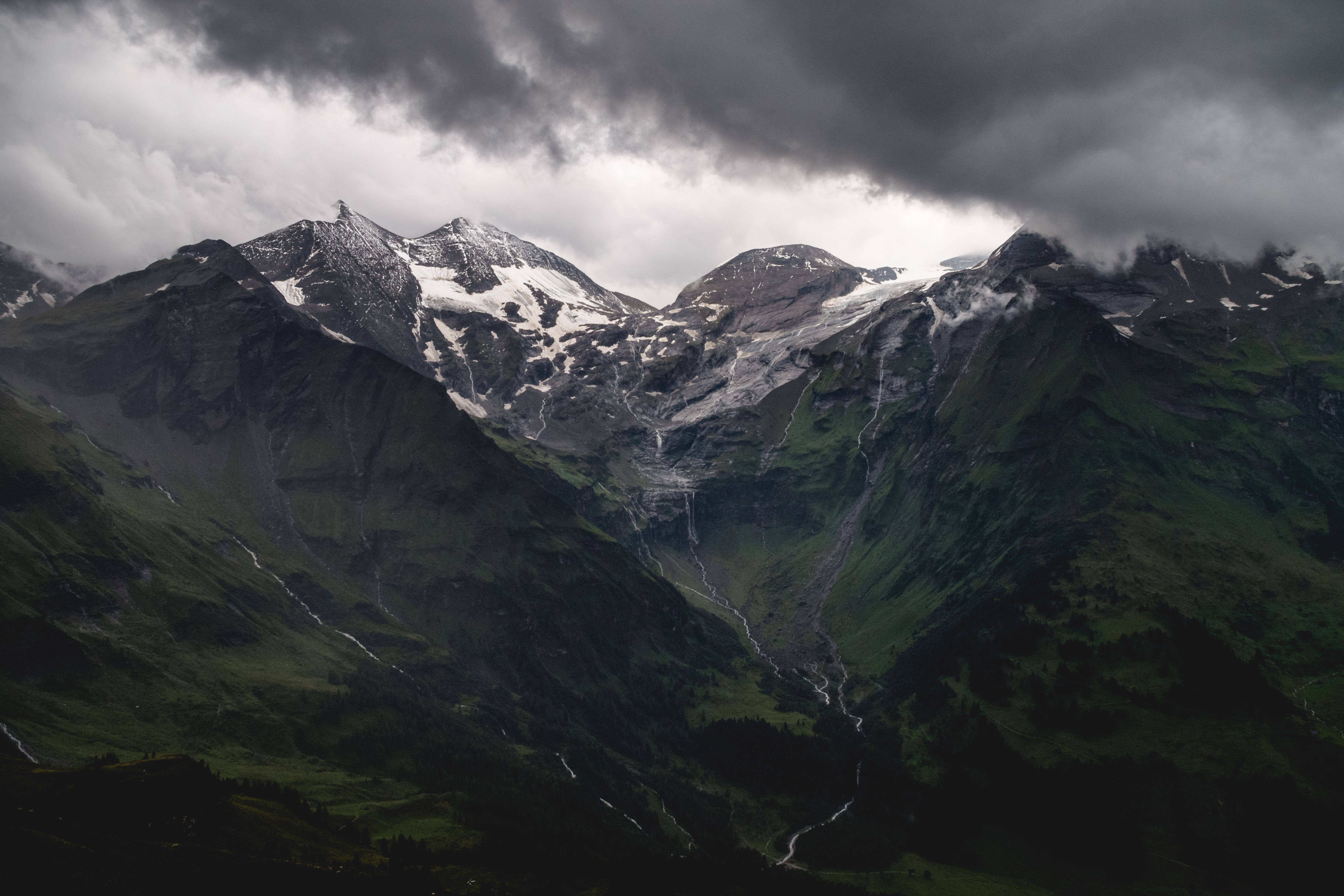 Dark 4K 桌布 壁紙 — 戲劇性的雪山峰在厚重的暴風雲下升起，擁有茂密的綠色山坡和層疊的瀑布。