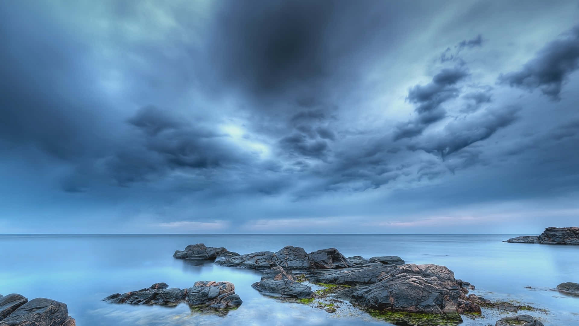 Calming Desktop 桌布 壁紙 — 戲劇性的風暴雲籠罩在平靜的海面上，前景是崎嶇的岩石。