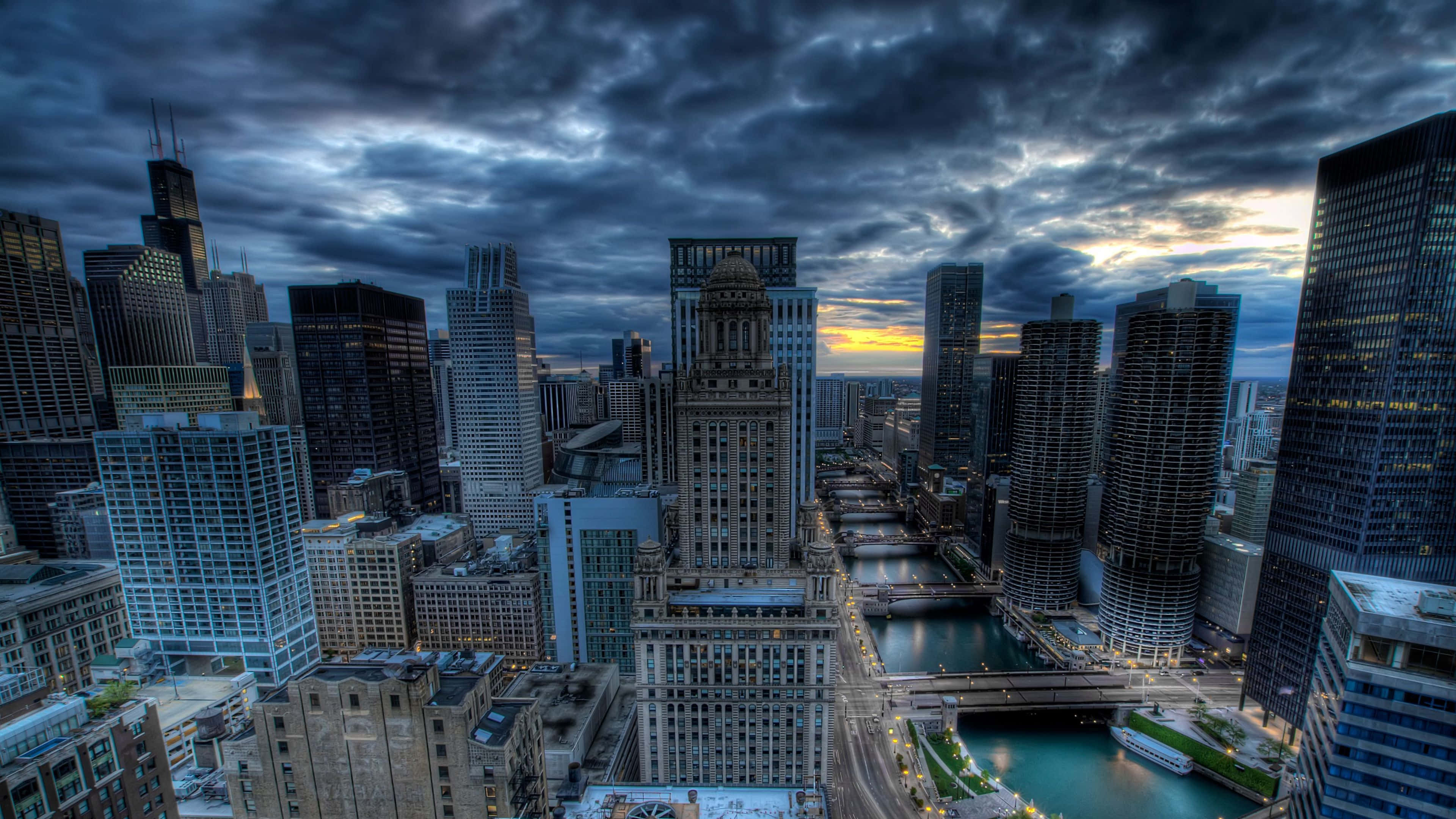 4K Ultra HD Chicago 桌布 壁紙 — 戲劇性的黃昏城市景觀，高聳的摩天大樓和蜿蜒穿過城市密集區的河流，在陰鬱的雲層下。