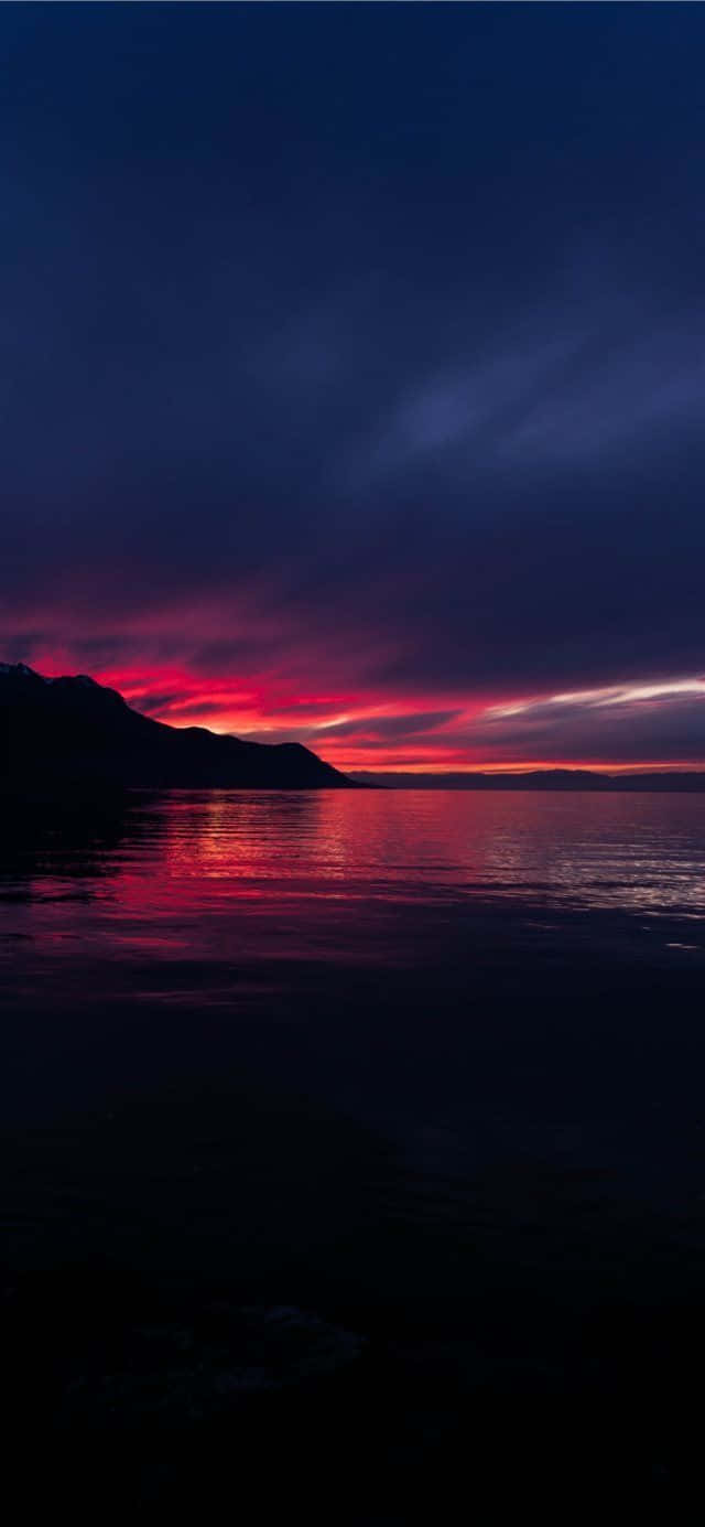 桌布 壁紙 — 戲劇性的黃昏天空，火紅和紫色的雲彩生動地倒映在平靜的水面上，旁邊是黑暗的山脈剪影。