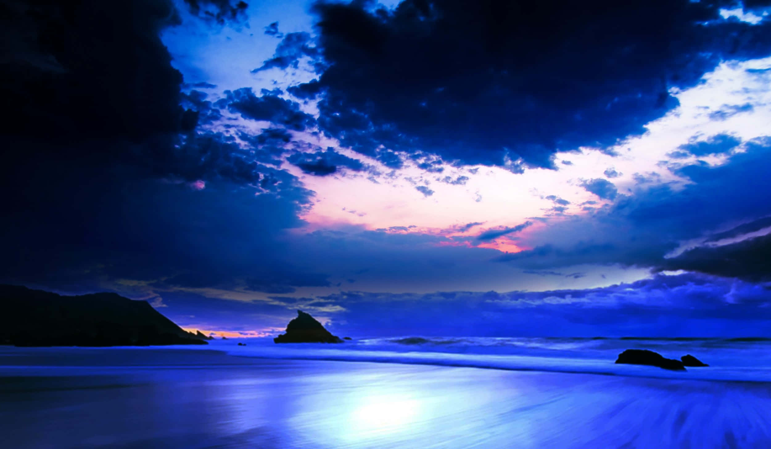 三星 桌布 壁紙 — 戲劇性的黃昏海灘場景，深藍色的海浪在風暴天空下拍打著黑暗的岩石，天空帶有粉紅色和紫色的色調。