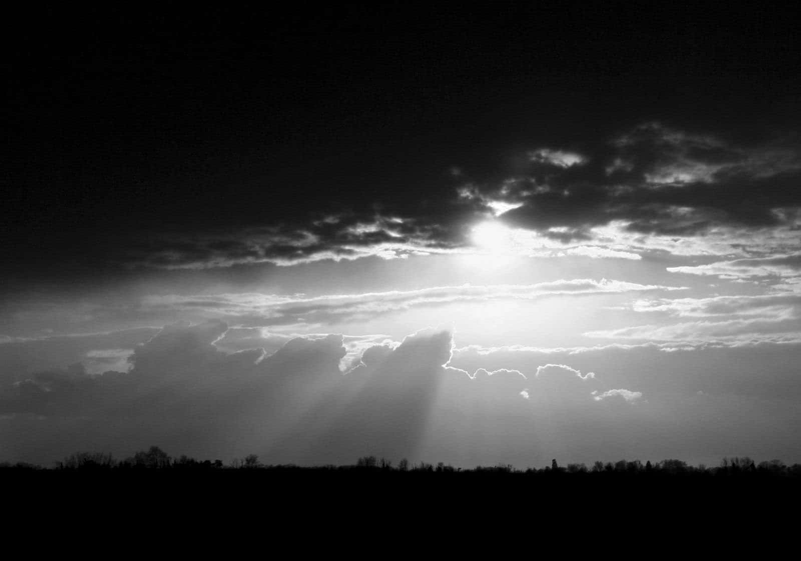 Black And White Sky 桌布 壁紙 — 戲劇性的黑白天空，陽光穿透烏雲，照射在剪影般的地平線上。