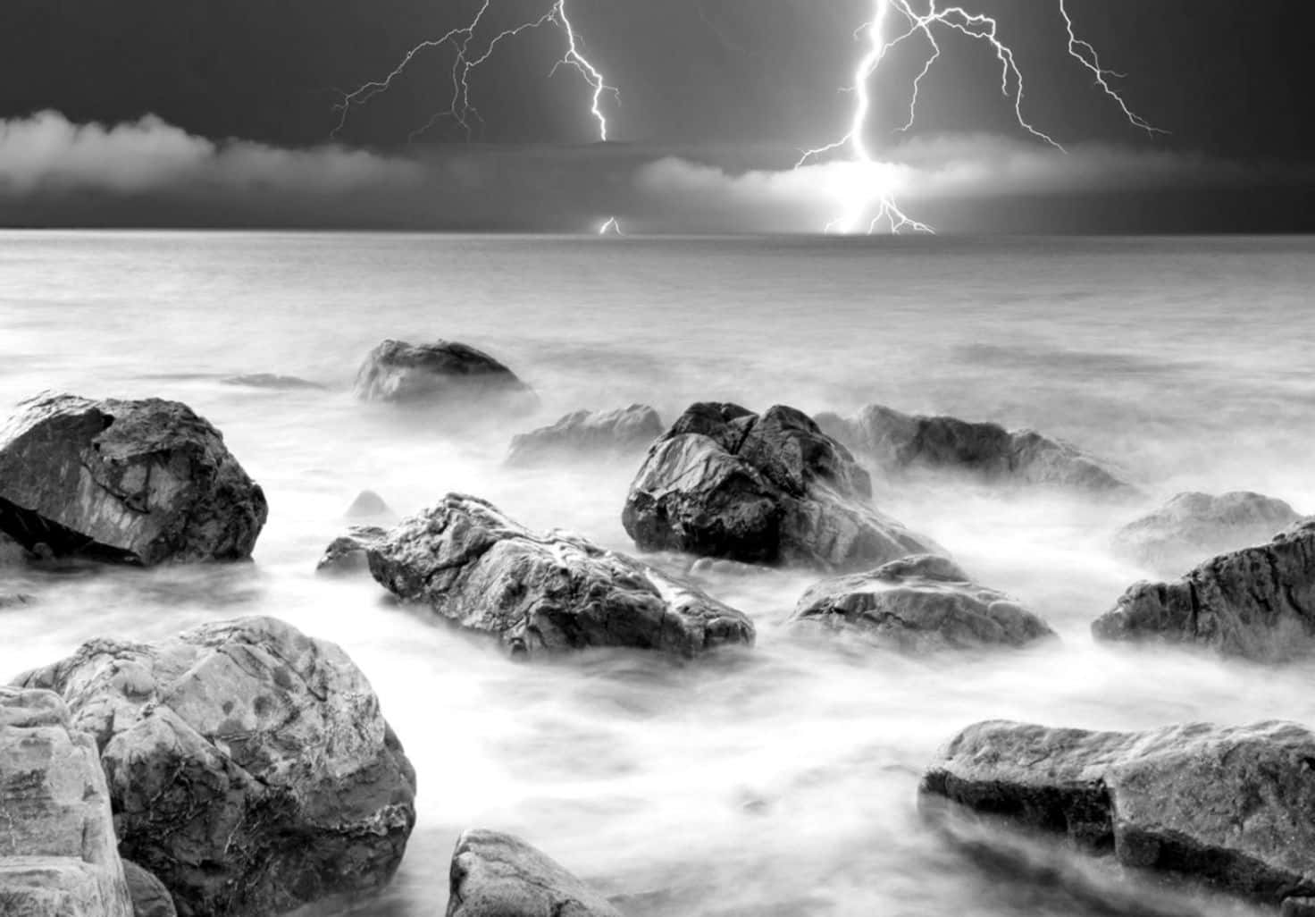 Black And White Storm 桌布 壁紙 — 戲劇性的黑白暴風雨場景，閃電劃過霧氣瀰漫的海洋，前景是崎嶇的岩石。