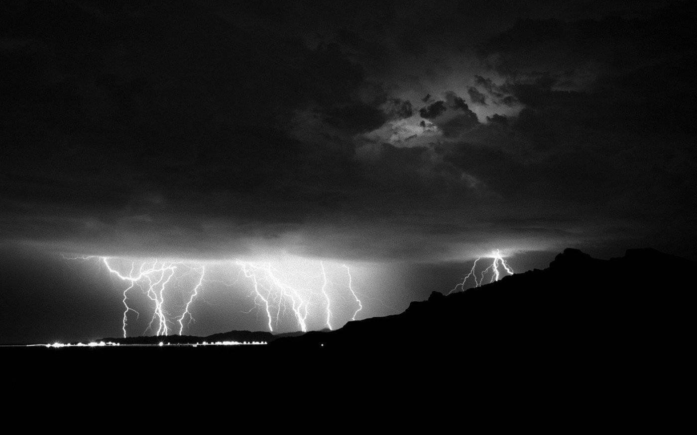 閃電 桌布 壁紙 — 戲劇性的黑白閃電照亮了暴風雨夜空中輪廓分明的山脈和遠處的城市燈光。