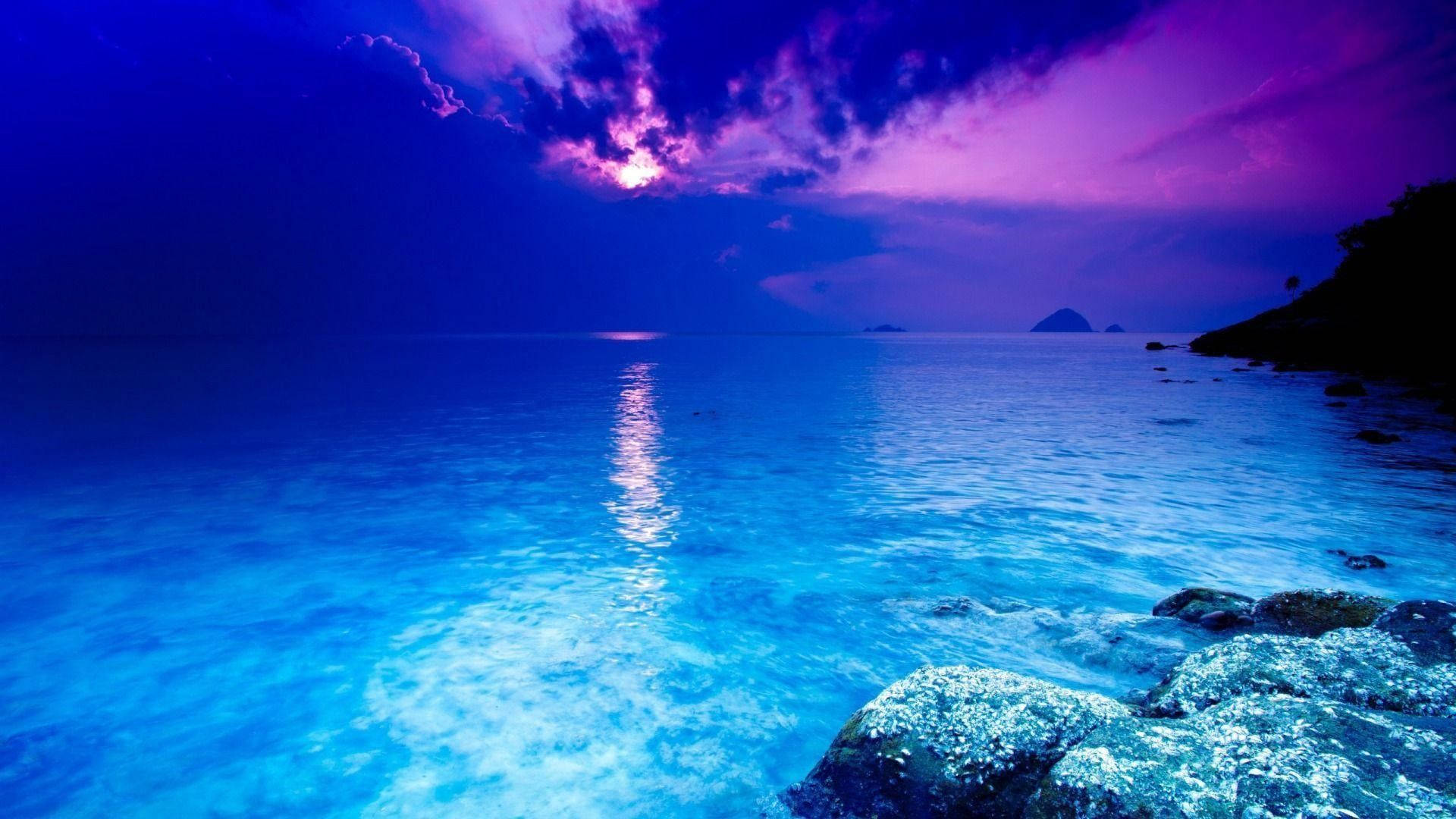 海洋 桌布 壁紙 — 戲劇性雲層下，平靜海洋上的鮮豔紫色和藍色日落，岩石海岸和遠處島嶼。