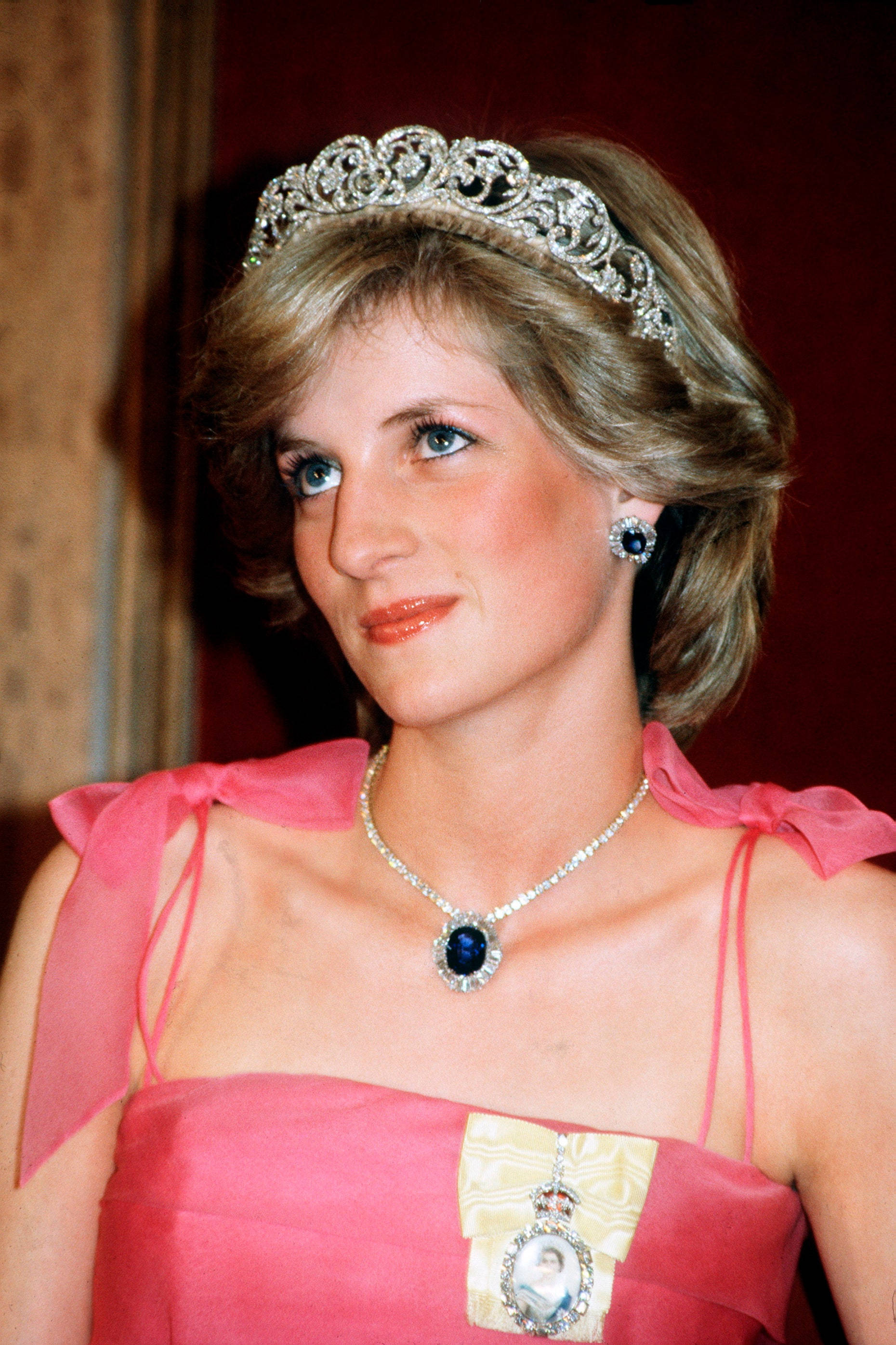Princess 桌布 壁紙 — 戴安娜王妃在濃郁的紅色背景下，身穿鮮豔的粉紅色禮服，佩戴閃閃發光的頭飾項鍊和相配的耳環，優雅地微笑。