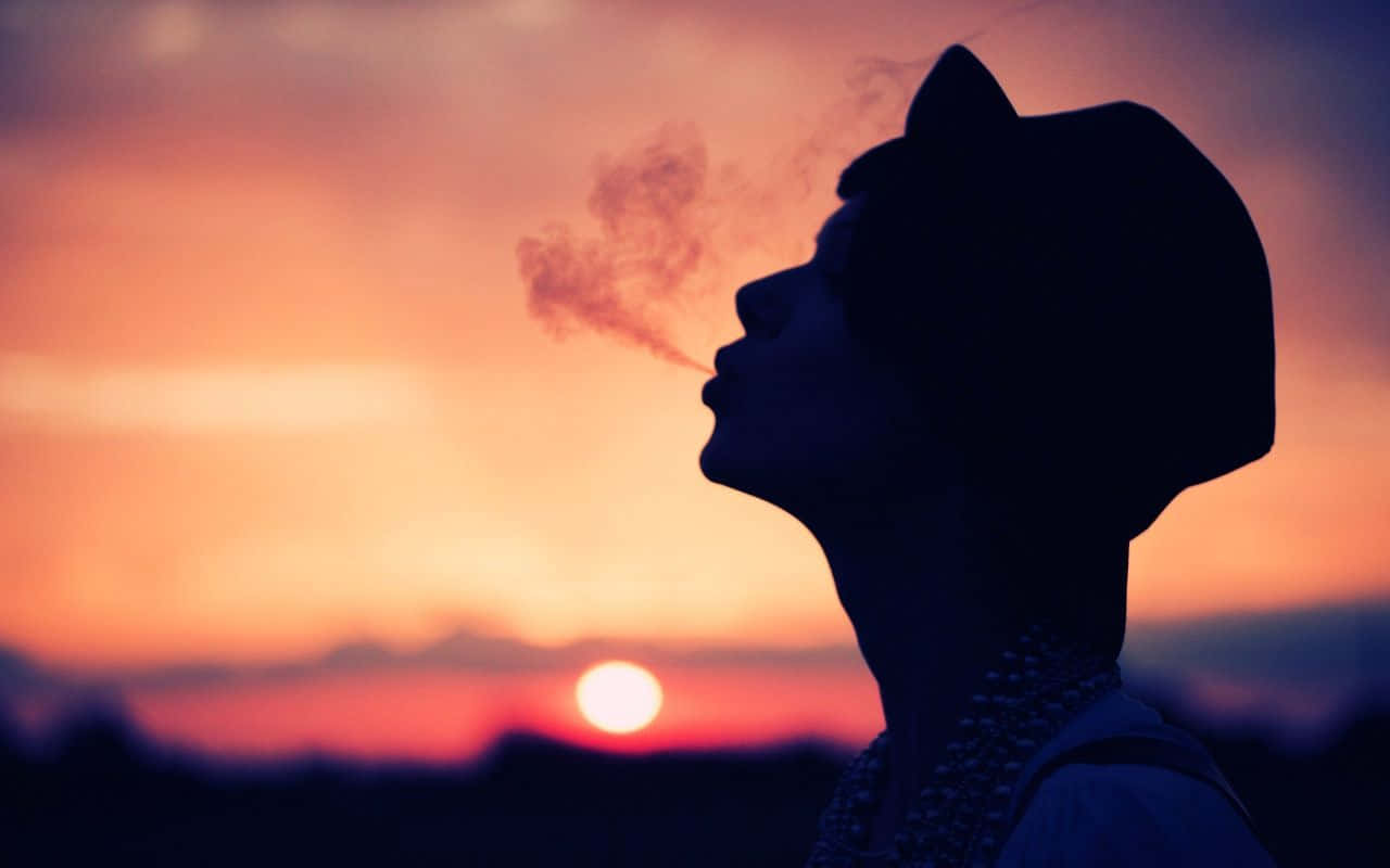 chill 桌布 壁紙 — 戴帽人物的剪影，在充滿活力的日落天空下呼出煙霧，背景有發光的太陽。