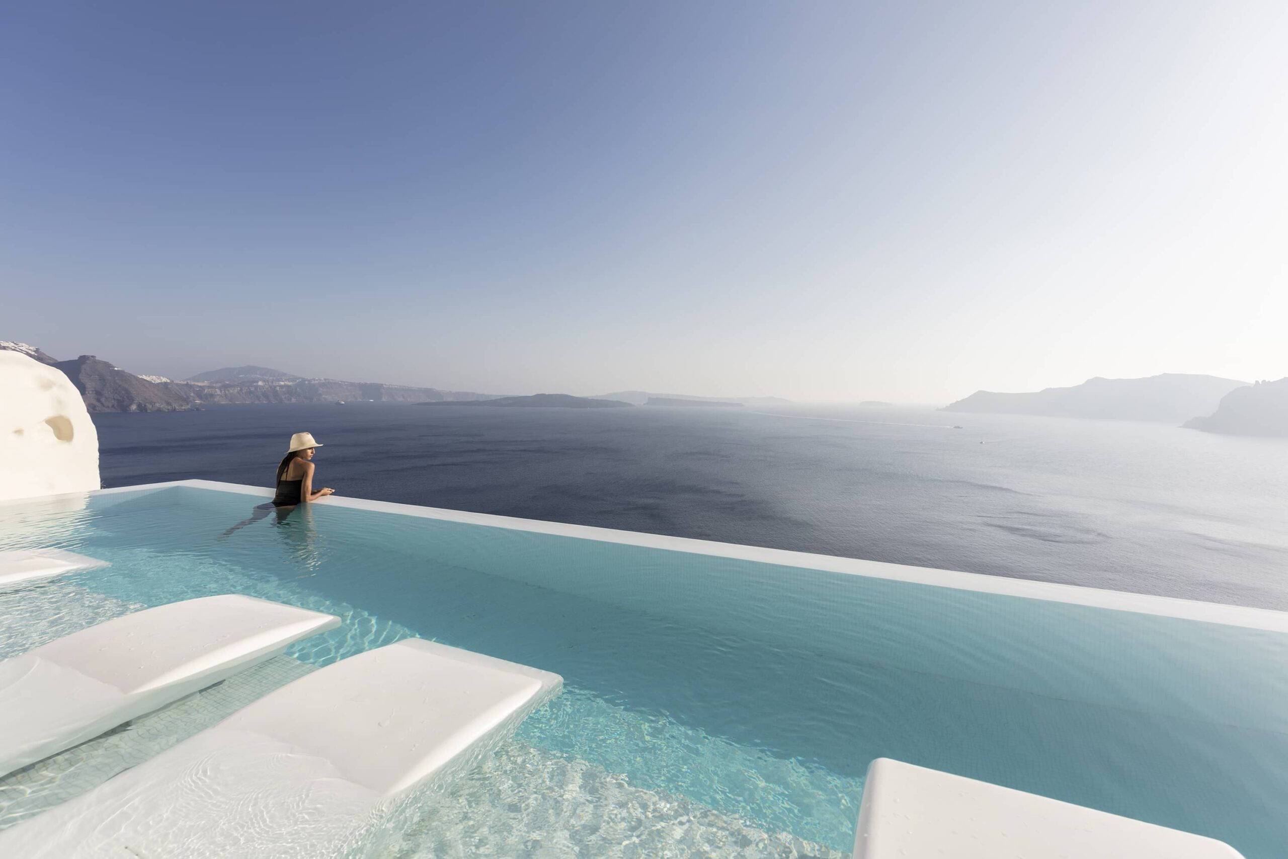 Santorini 桌布 壁紙 — 戴帽子的人正在白色無邊際泳池中放鬆，俯瞰深藍色的愛琴海和火山懸崖，天空晴朗。