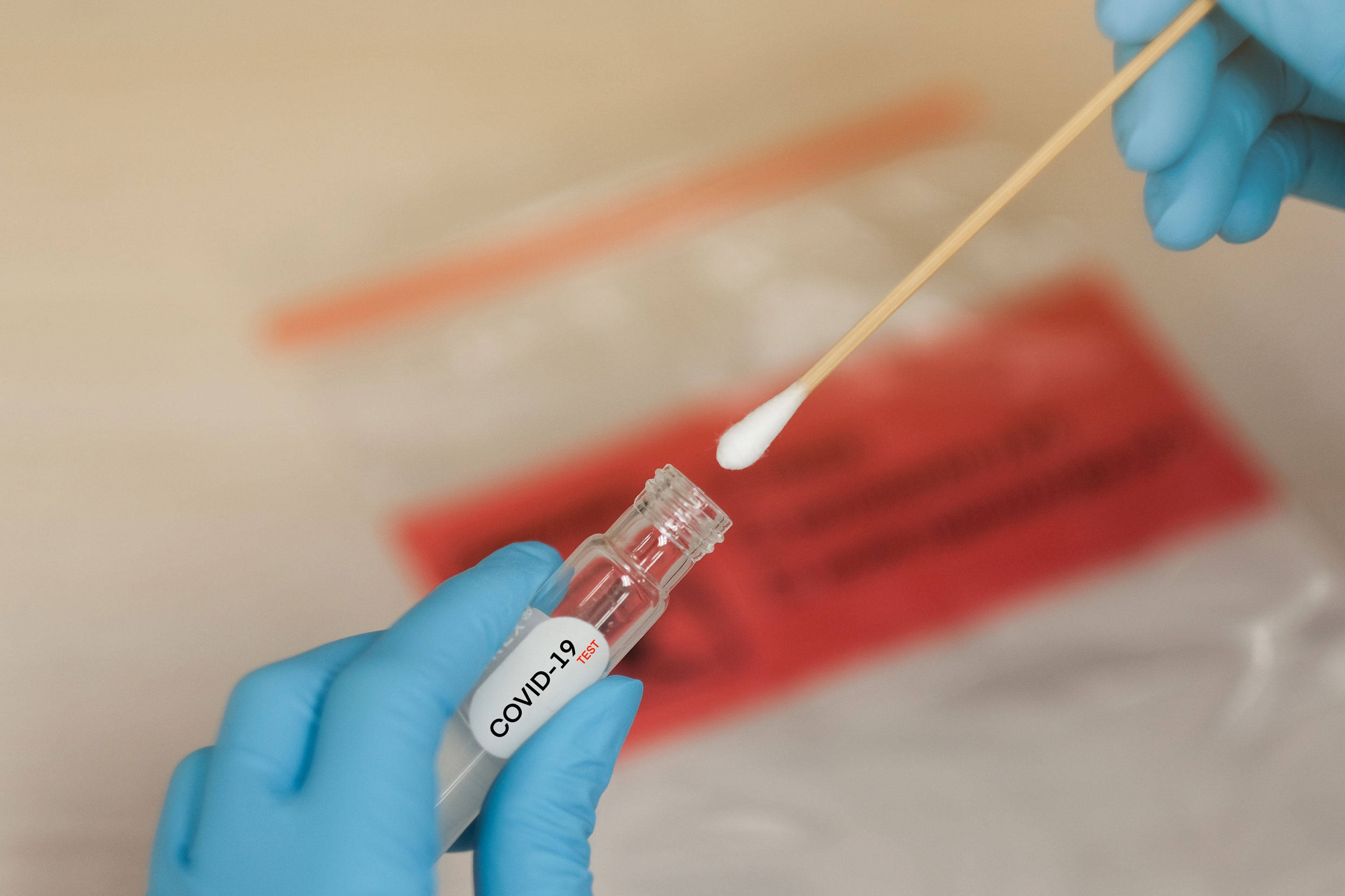 Covid 桌布 壁紙 — 戴手套的手小心地將拭子插入標有標籤的COVID-19測試瓶，背景為模糊的紅色和米色。