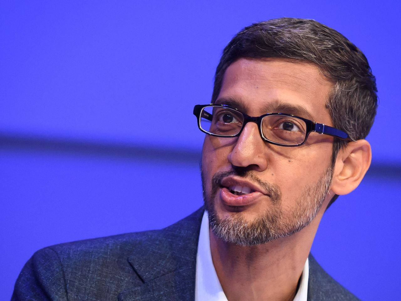 google 桌布 壁紙 — 戴眼鏡和鬍鬚的男子在純藍色背景前說話的特寫肖像。