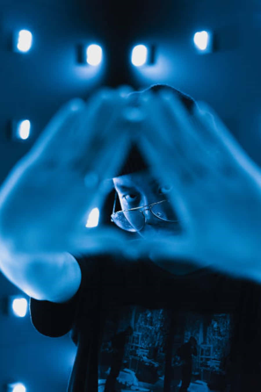 illuminati 桌布 壁紙 — 戴眼鏡的人用雙手形成三角形框住臉龐，在冷色藍色舞台燈光下。