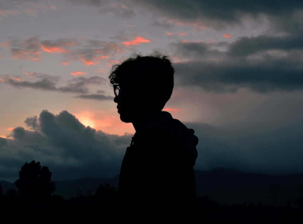 Silhouette 桌布 壁紙 — 戴眼鏡的人的剪影，凝視著壯麗的日落天空，有橙色雲和暗雲。
