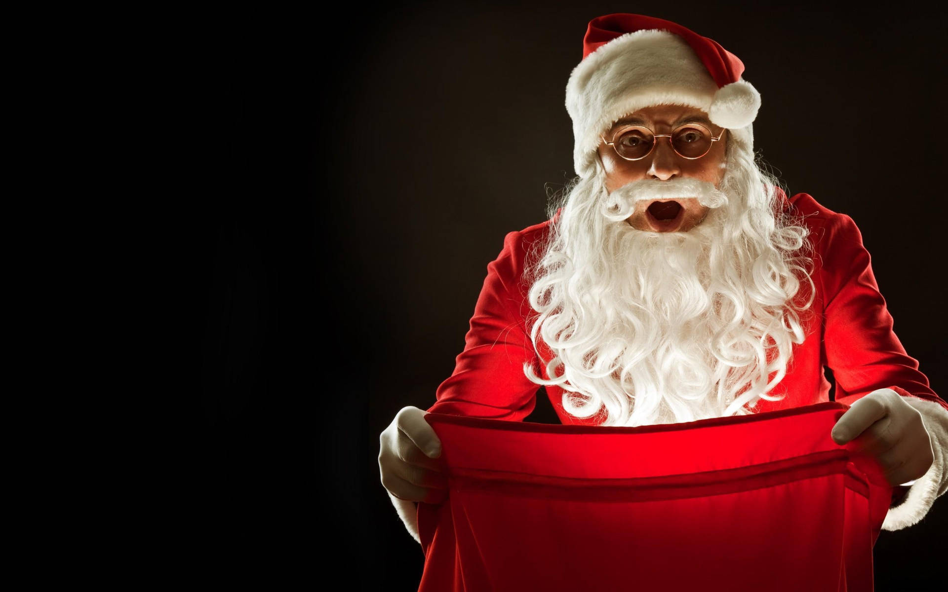 Santa Claus 桌布 壁紙 — 戴眼鏡的聖誕老人帶著驚訝的表情，在深色背景前打開一個紅色袋子。