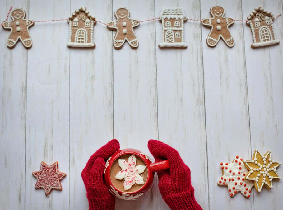 Cozy Winter Desktop 桌布 壁紙 — 戴紅色手套的雙手捧著一杯熱可可，上面漂浮著雪花餅乾，周圍是薑餅餅乾，放在白色木質表面上。