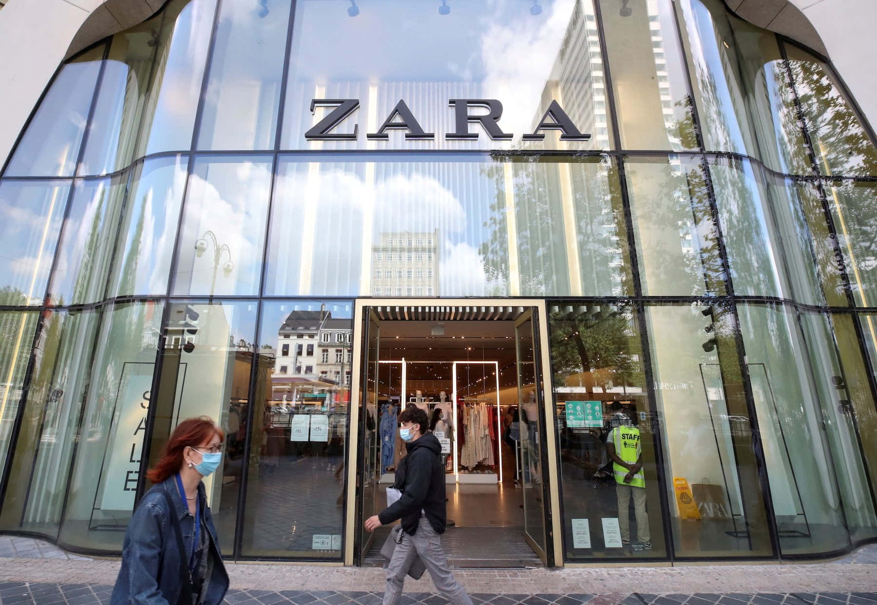 Zara 桌布 壁紙 — 戴著口罩的人們走過一間現代化的玻璃幕牆Zara商店，倒映著明亮的天空和城市建築。