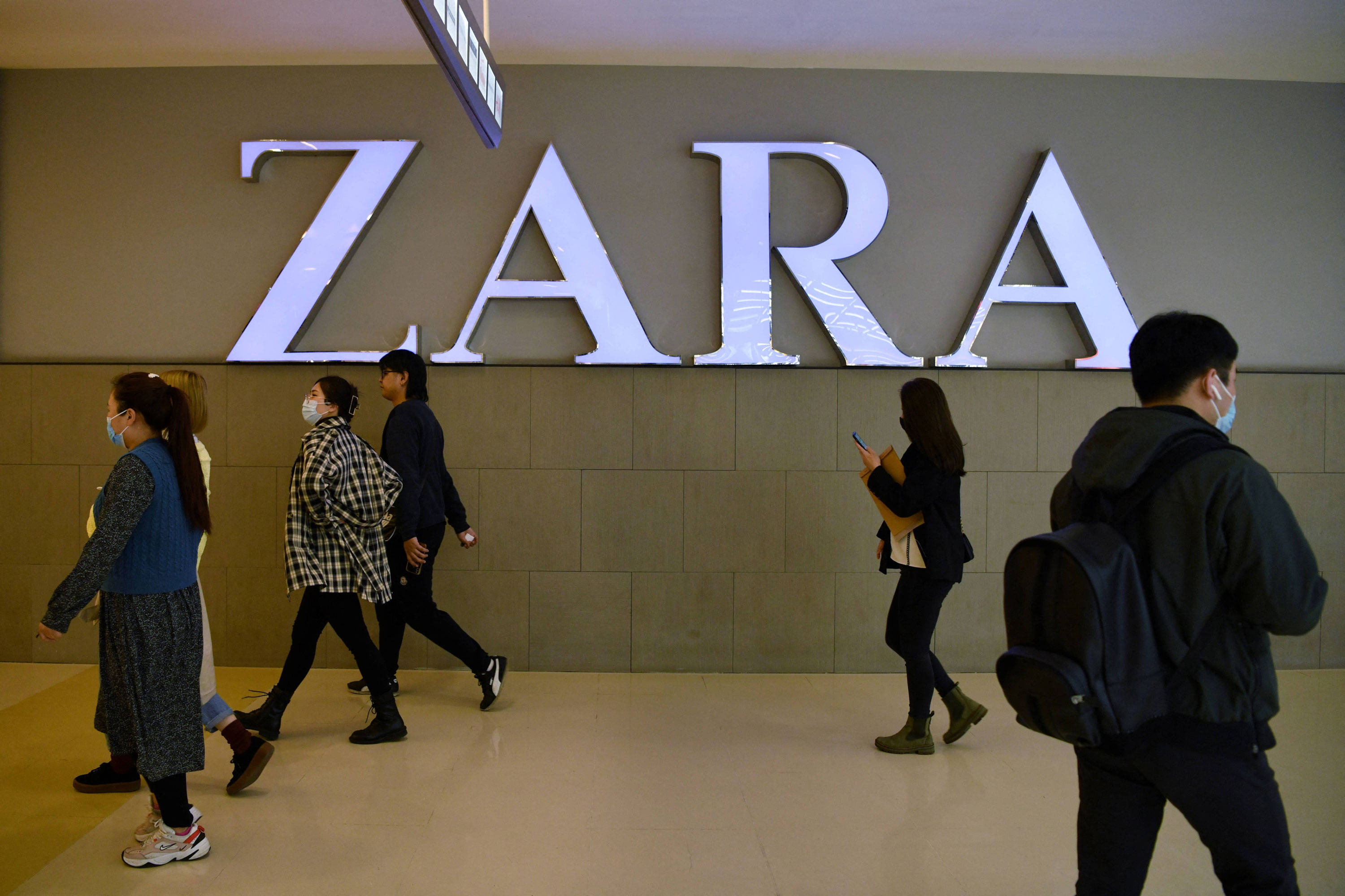 Zara 桌布 壁紙 — 戴著口罩的人們走過現代購物中心內部一個大型發光的ZARA商店標誌。
