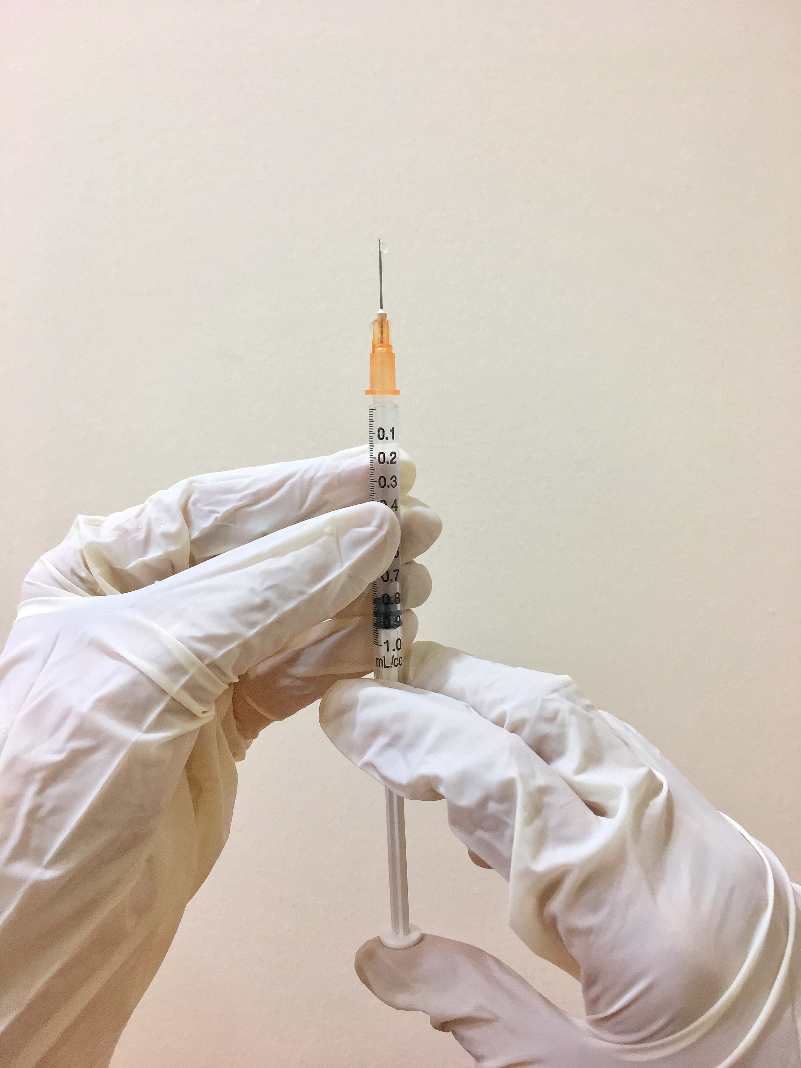 生病 桌布 壁紙 — 戴著手套的手握著一支醫療注射器，針頭準備好注射，背景是純色米色牆壁。