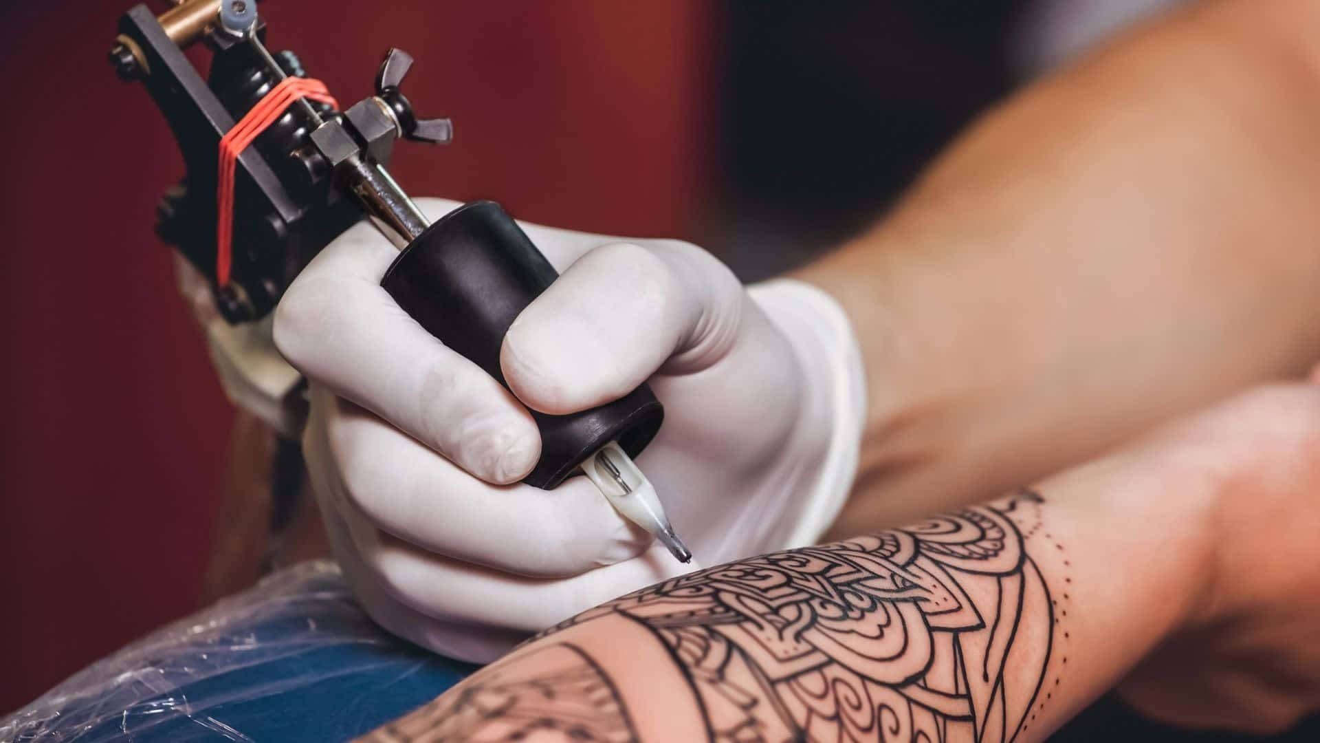 Tattoo 桌布 壁紙 — 戴著手套的手握著紋身機，在人的前臂上蝕刻出複雜的黑色曼陀羅圖案。