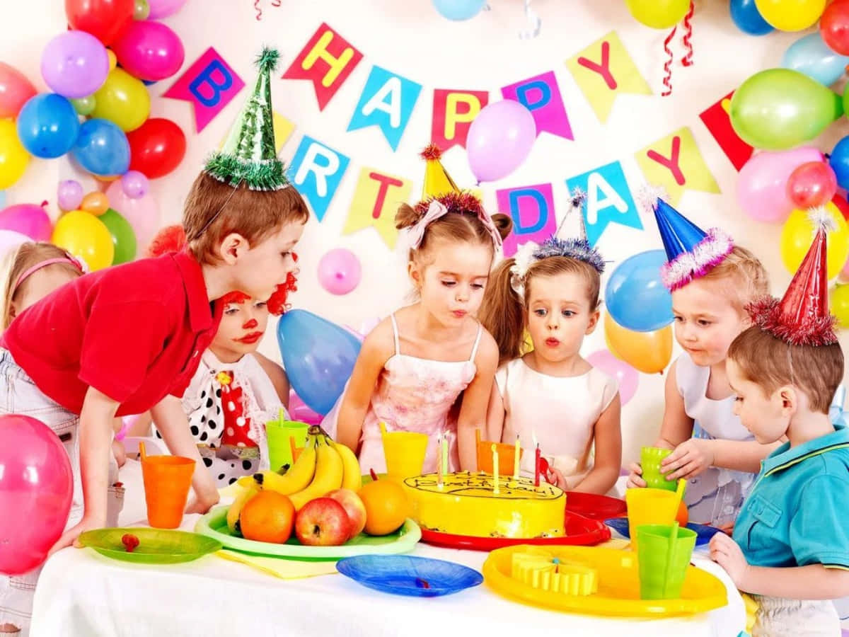 Birthday Celebration 桌布 壁紙 — 戴著派對帽的孩子們聚集在生日蛋糕周圍，蛋糕上有蠟燭和水果，上方有彩色氣球和「生日快樂」橫幅。