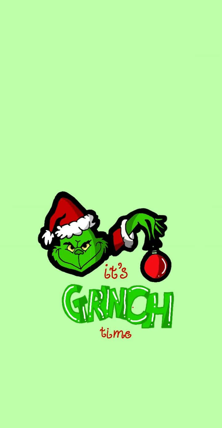 Cute Grinch 桌布 壁紙 — 戴著紅色聖誕帽的鬼靈精在淺綠色背景前拿著一個紅色裝飾，上面有文字 it's GRINCH time。